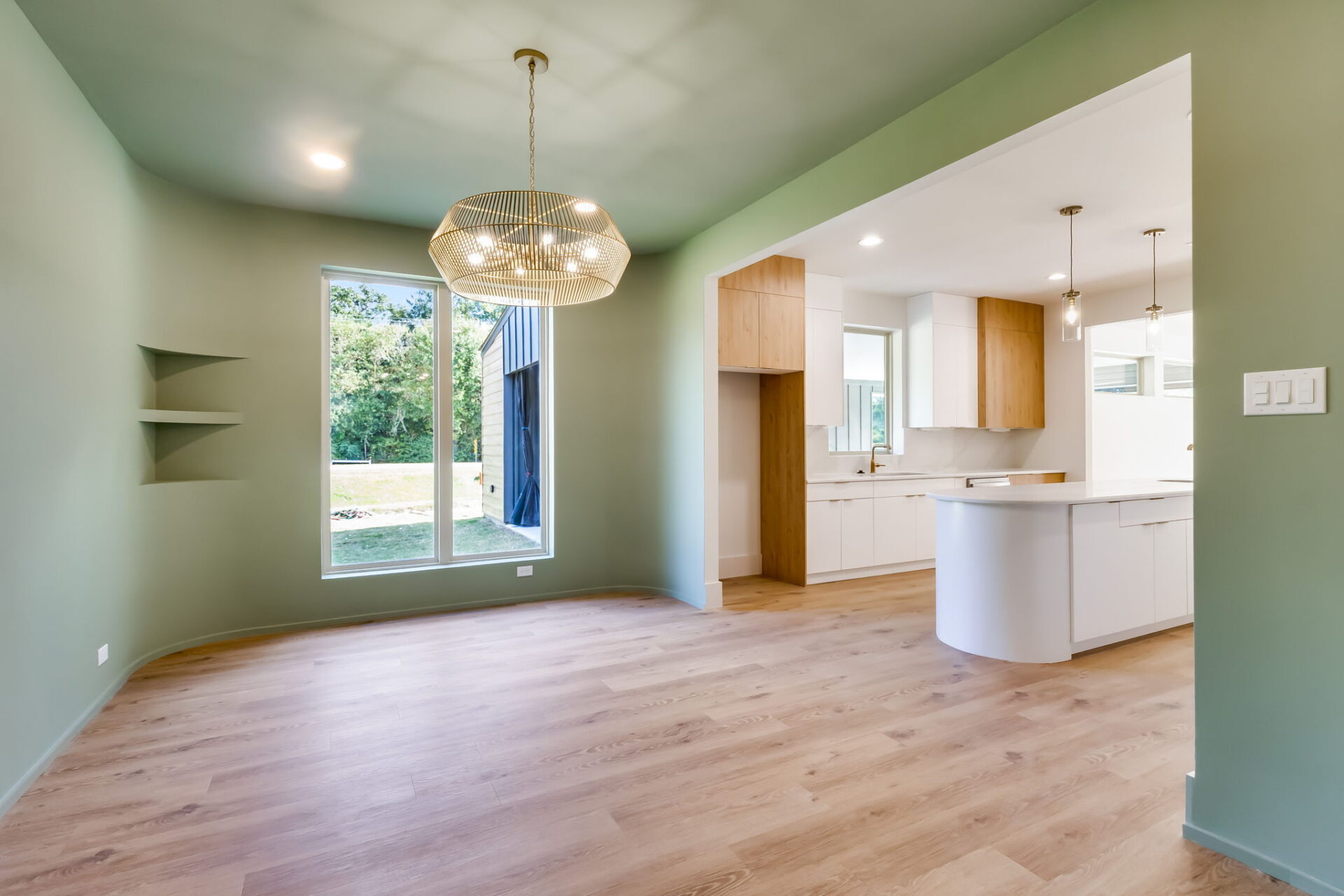 A modern dining area with green walls, light wood flooring, a large window, built-in shelves, and a round pendant light, adjacent to a white kitchen with wood cabinetry and an island.