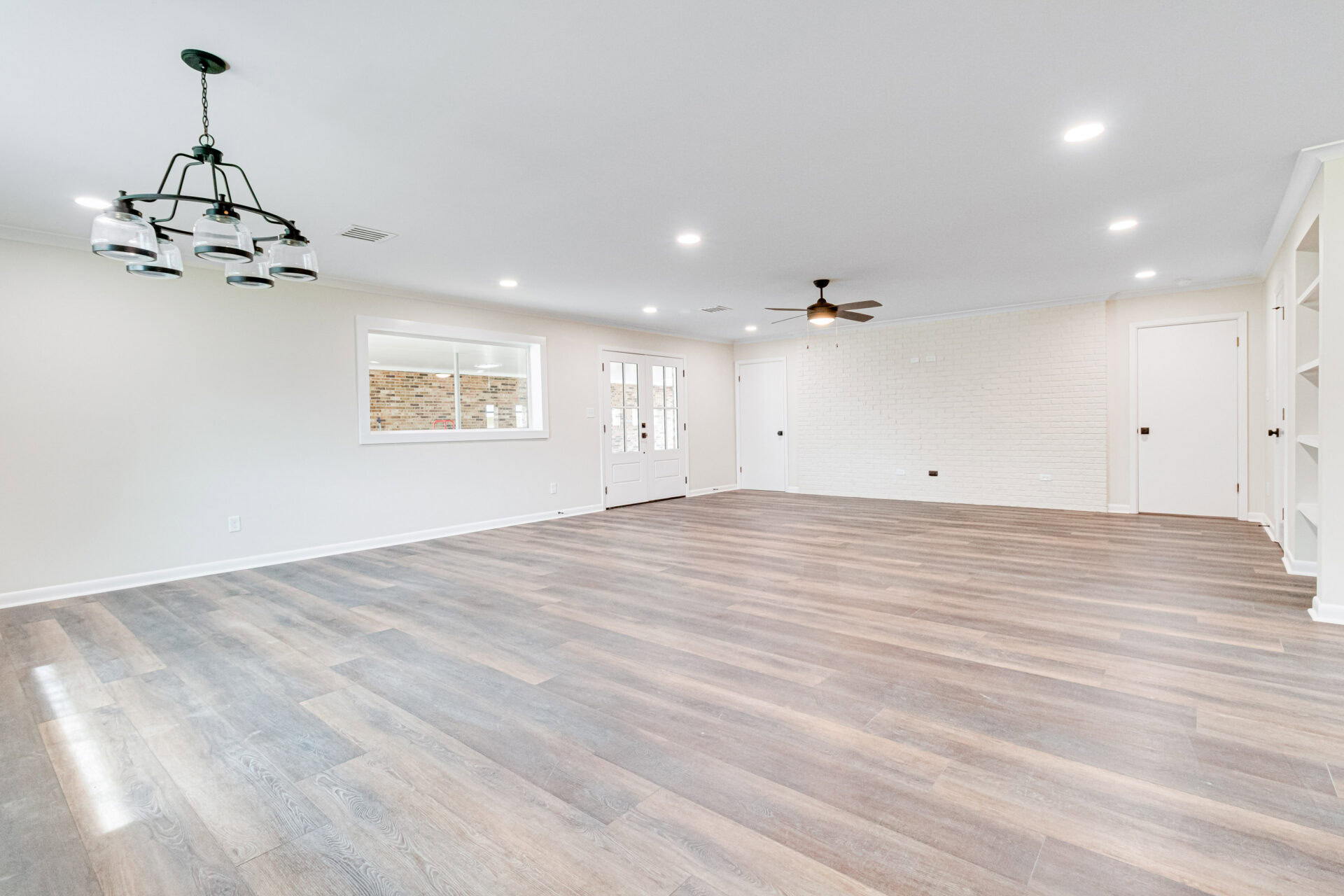 Spacious empty room with wood flooring, white walls, recessed lighting, a ceiling fan, a decorative light fixture, a window, a glass-paneled door, and built-in shelves in the corner.