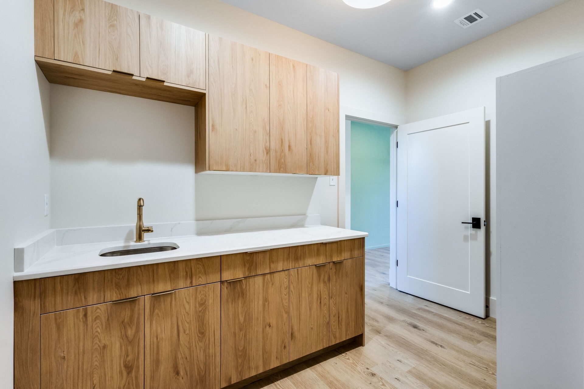 A modern laundry room with light wood cabinets, a white countertop, an undermount sink with a gold faucet, and a white door open to an adjacent room. The floor is light wood, and the walls are off-white.
