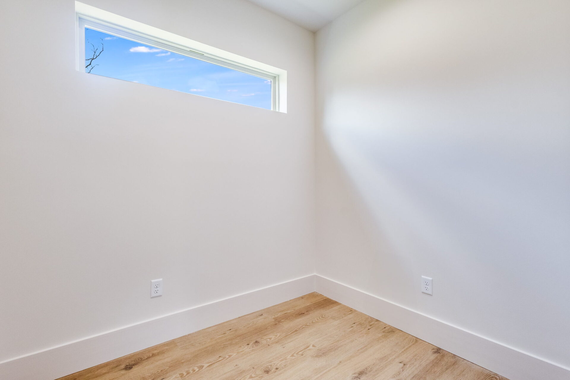 A small, empty room with white walls, light wood flooring, and a narrow horizontal window near the ceiling letting in natural light. Two electrical outlets are visible on the lower part of the walls.