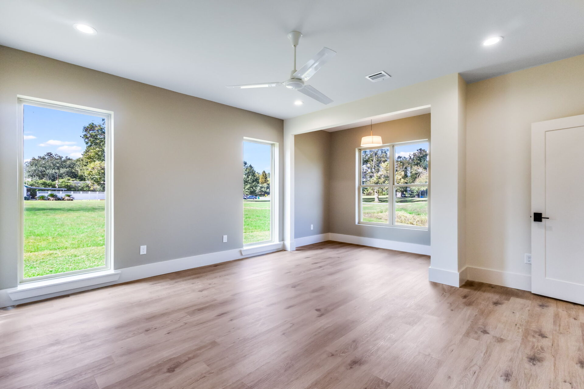A spacious, empty room with light wood flooring, beige walls, large windows letting in natural light, a ceiling fan, and a small alcove area with a hanging light fixture. Green grass and trees are visible outside.