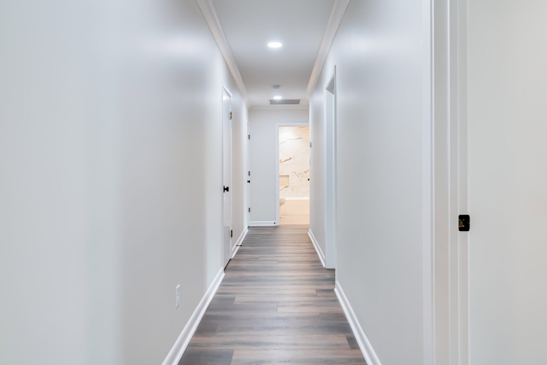 A long, narrow hallway with white walls and a wood-patterned floor, illuminated by recessed ceiling lights, leading to a brightly lit bathroom with marble walls at the end.