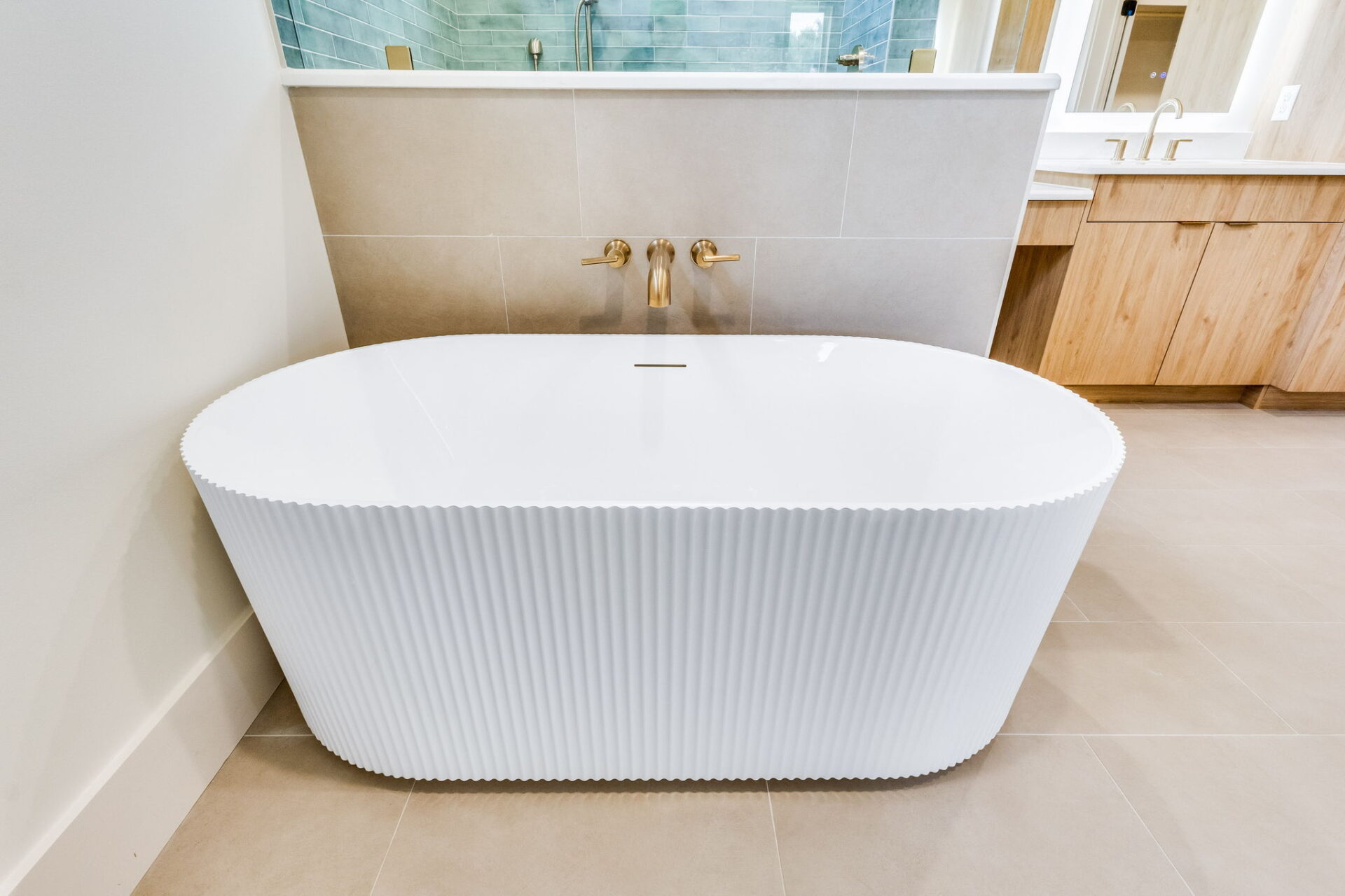 A modern bathroom with a white freestanding bathtub featuring a textured exterior, gold wall-mounted faucet, beige tile floor, and a wooden vanity with dual sinks and gold fixtures in the background.