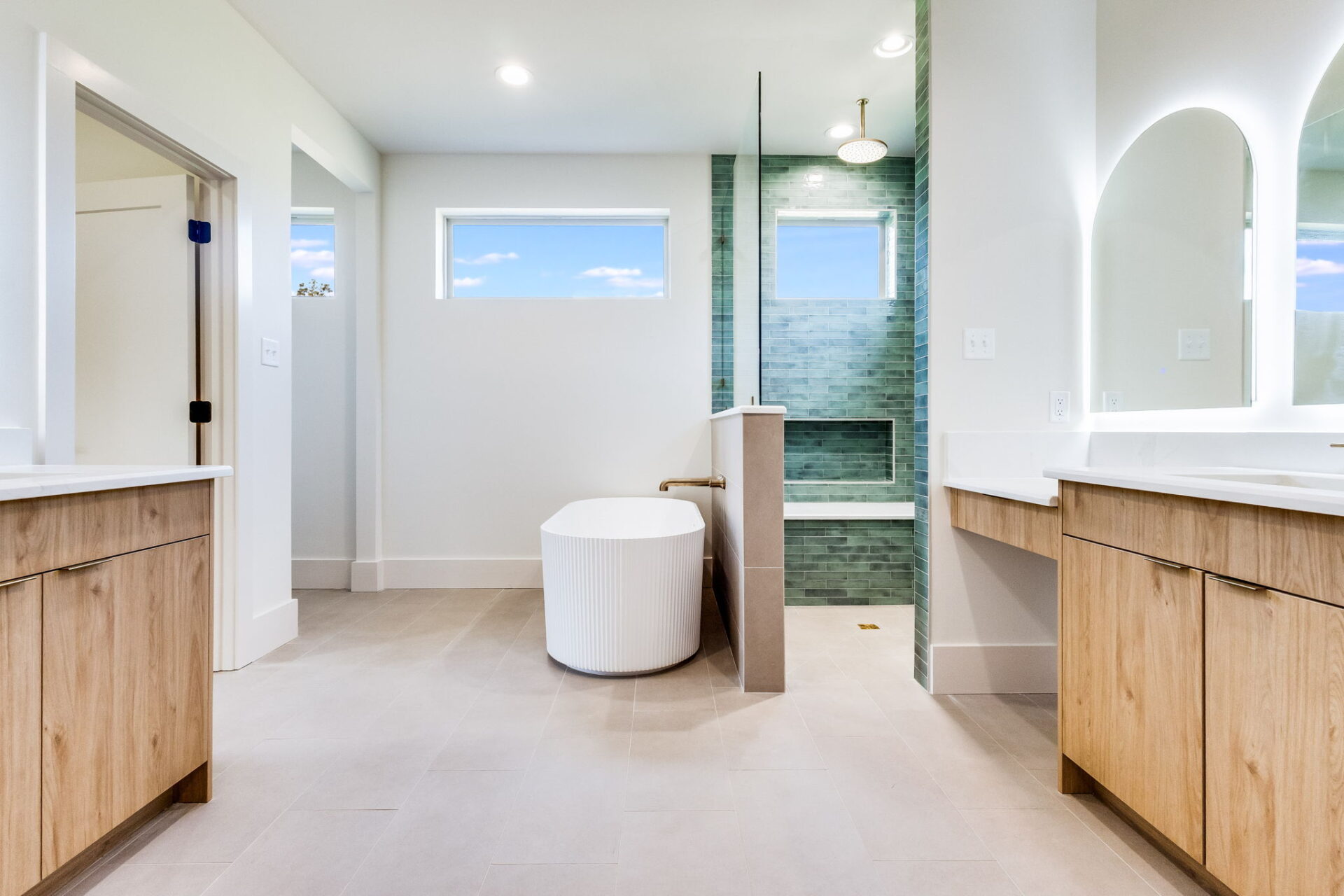 Modern bathroom with light wood cabinets, a freestanding white bathtub, a walk-in shower with green tile, large mirrors with built-in lighting, and a window letting in natural light.