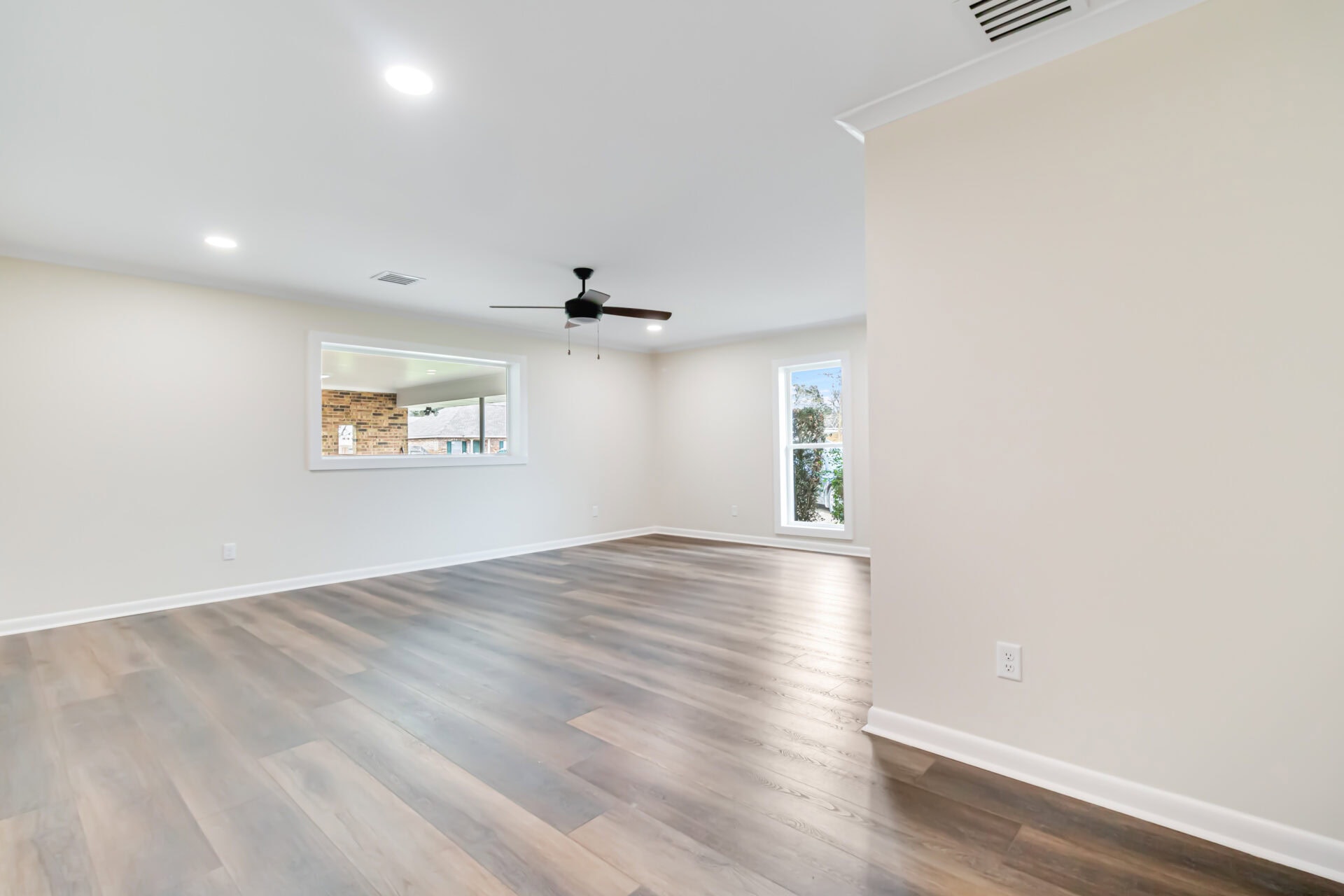A spacious, empty room with light-colored walls, wood-look flooring, recessed ceiling lights, a ceiling fan, a large window, and an interior window showing a brick feature in the adjacent space.