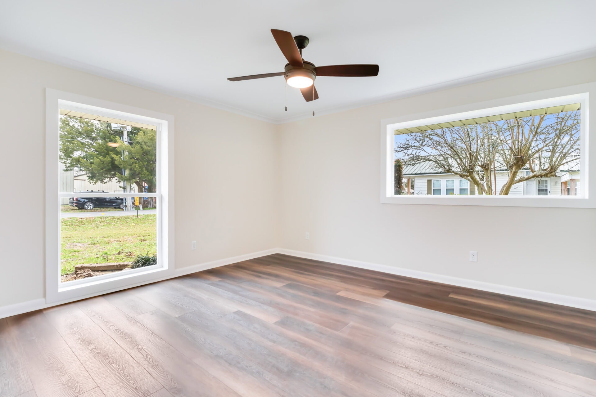 Empty room with light-colored walls, two large windows showing a yard and houses outside, dark wood flooring, and a ceiling fan with a light in the center of the ceiling.