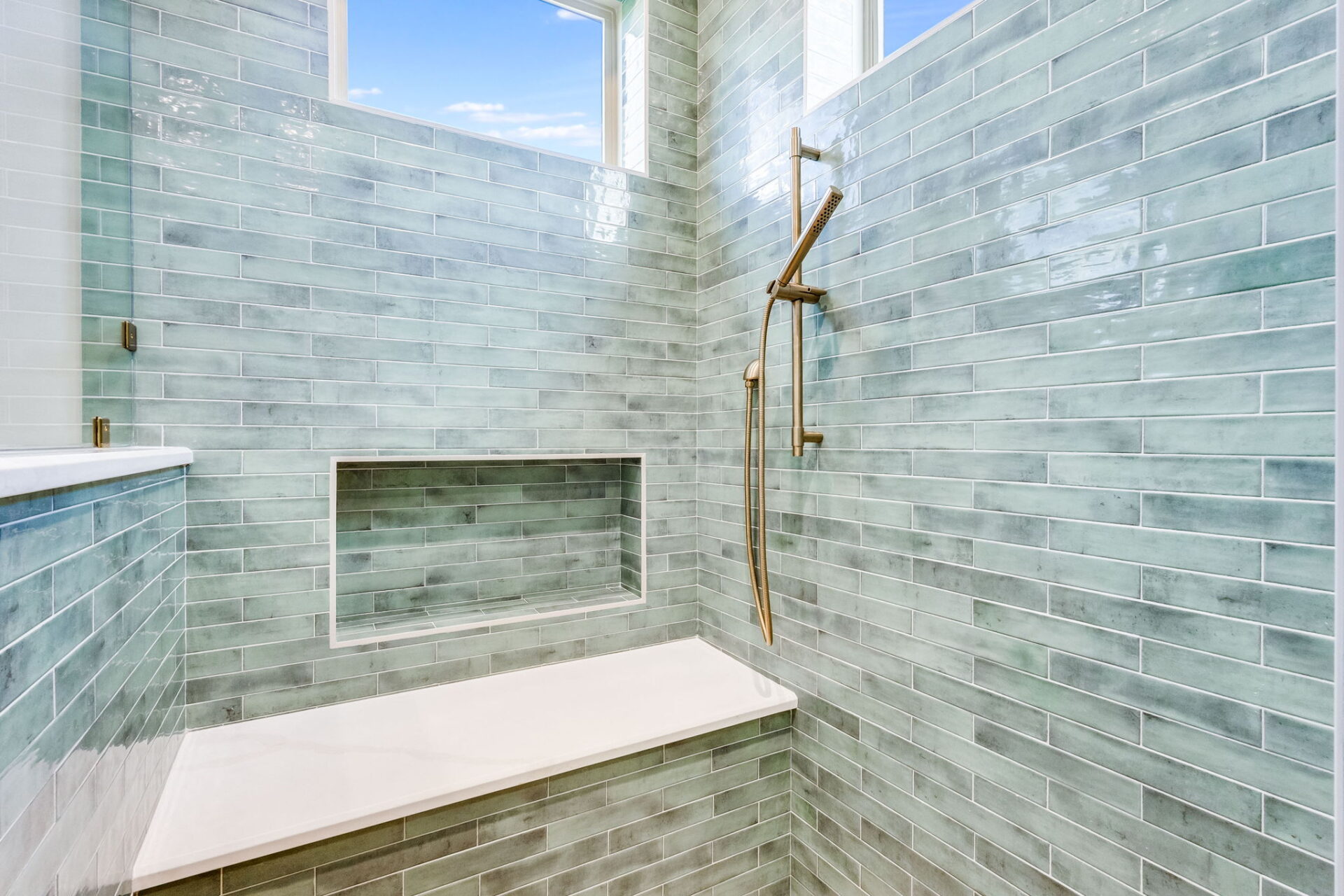 A shower with light blue-green subway tiles, a wall-mounted gold showerhead and handle, a built-in shelf, a white bench, and two windows letting in natural light.
