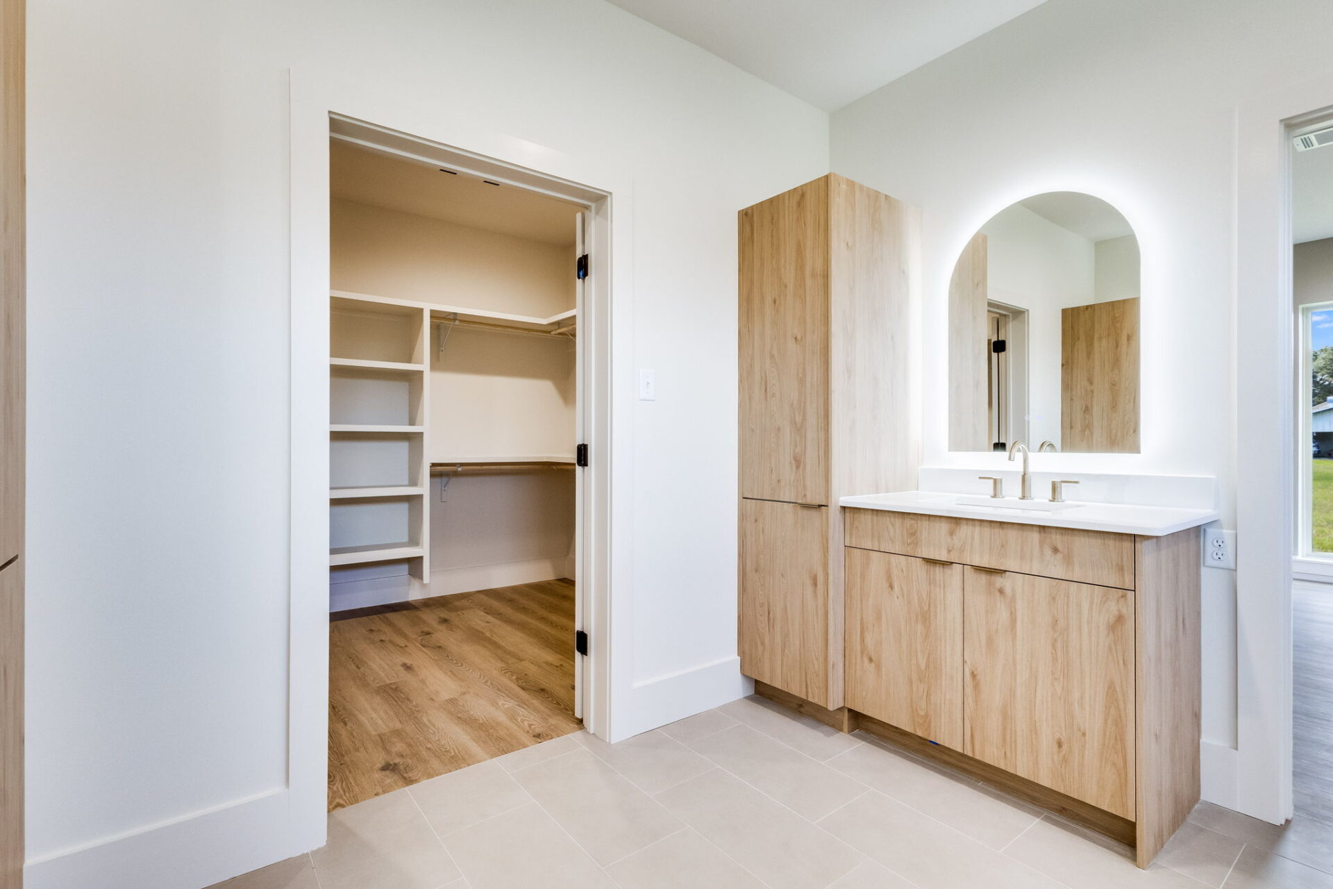 A bathroom with a light wood vanity, white countertop, arched mirror, and built-in sink. An open doorway leads to a walk-in closet with shelves and wood flooring. The room has white walls and tile flooring.
