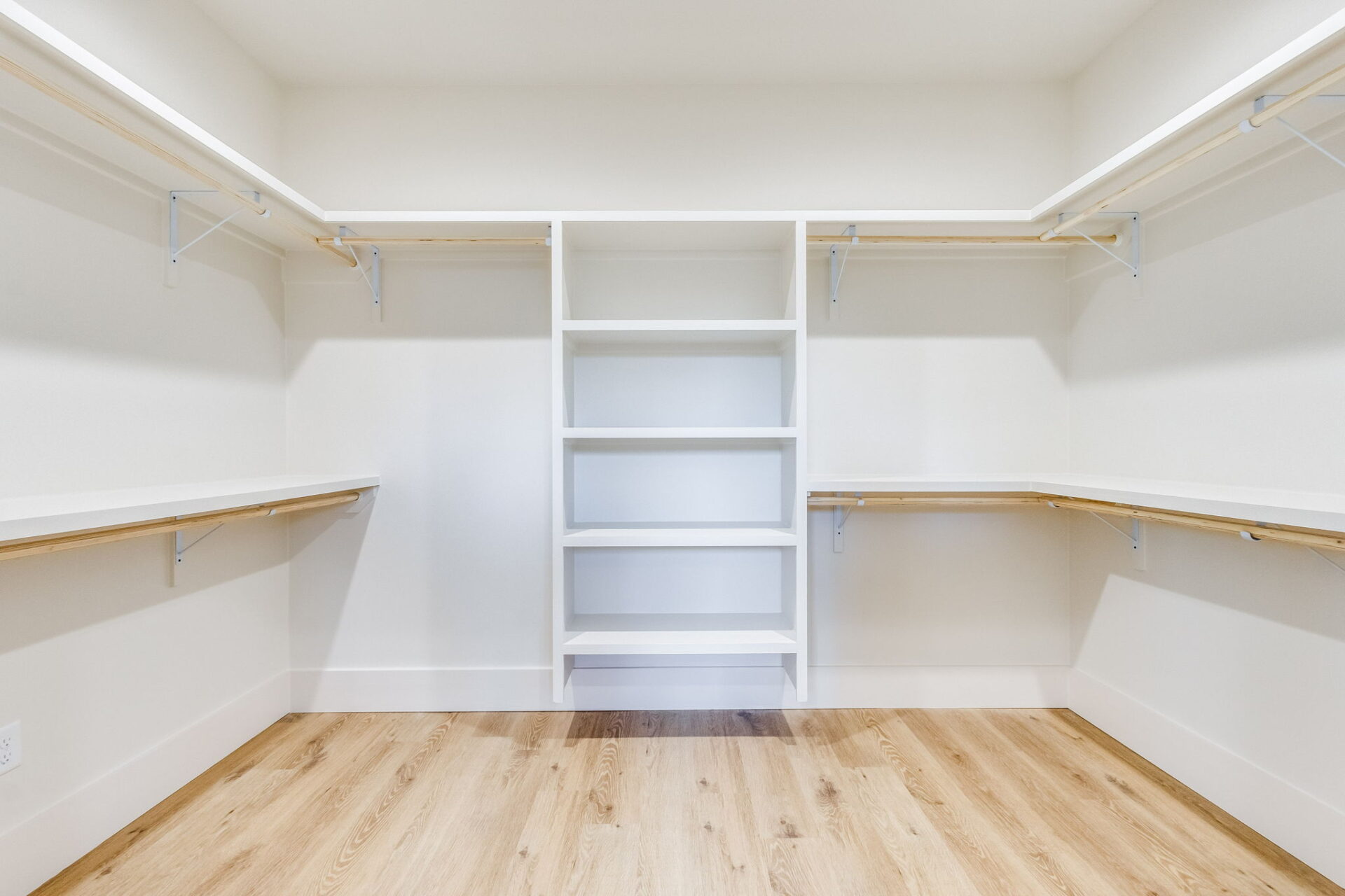 Empty walk-in closet with light wood flooring, white walls, open shelving unit in the center, and wooden hanging rods with white supports on both sides of the room. The closet is well-lit and spacious.