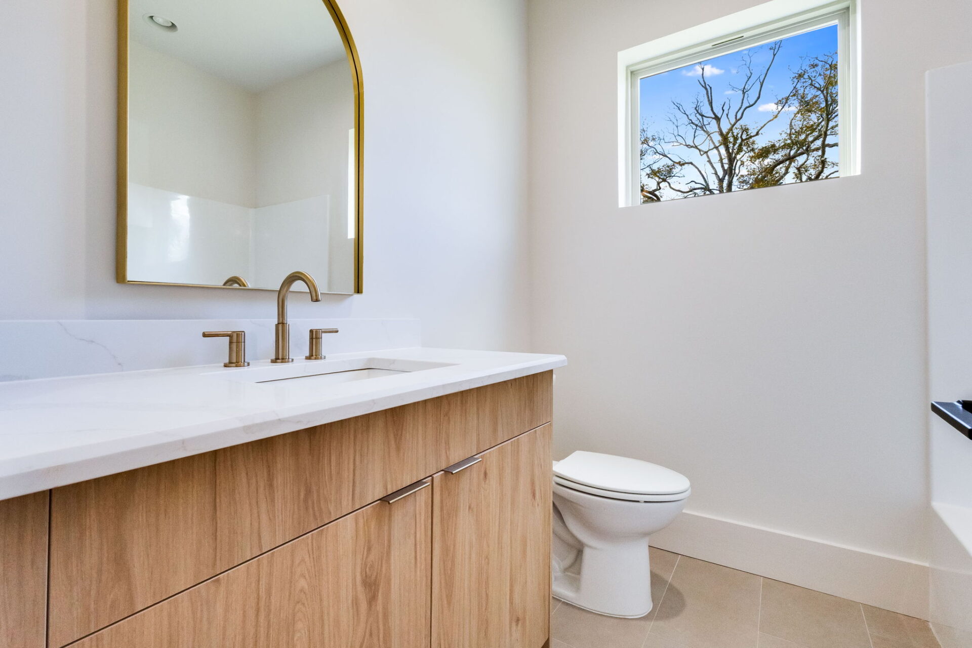 A modern bathroom with a light wood vanity, white countertop, dual faucets, a rectangular mirror with a gold frame, a white toilet, a small window, and neutral-colored tile flooring.