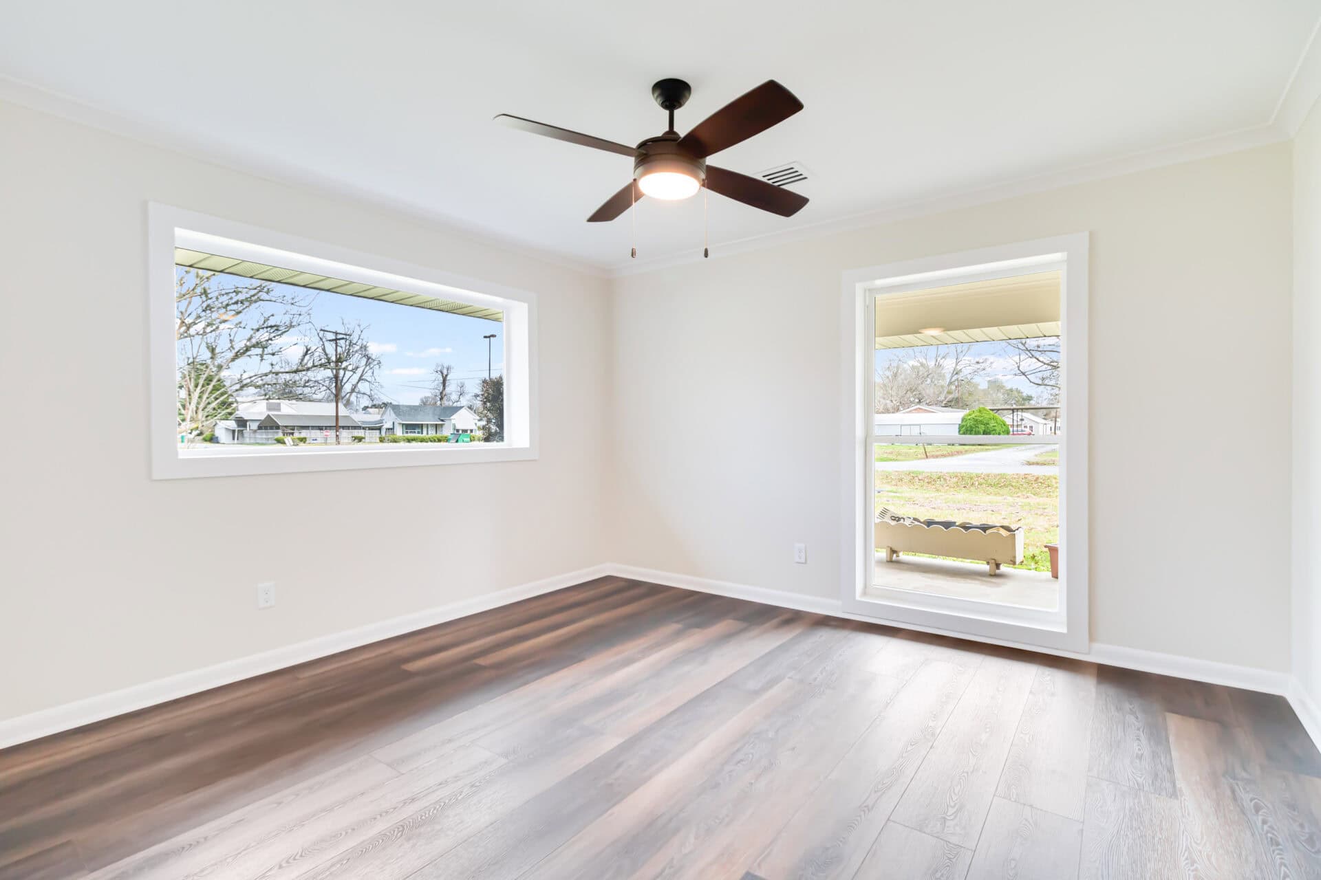 Empty room with light-colored walls, dark wood flooring, a ceiling fan with lights, and two large windows allowing natural light to enter. The windows show a view of trees and houses outside.