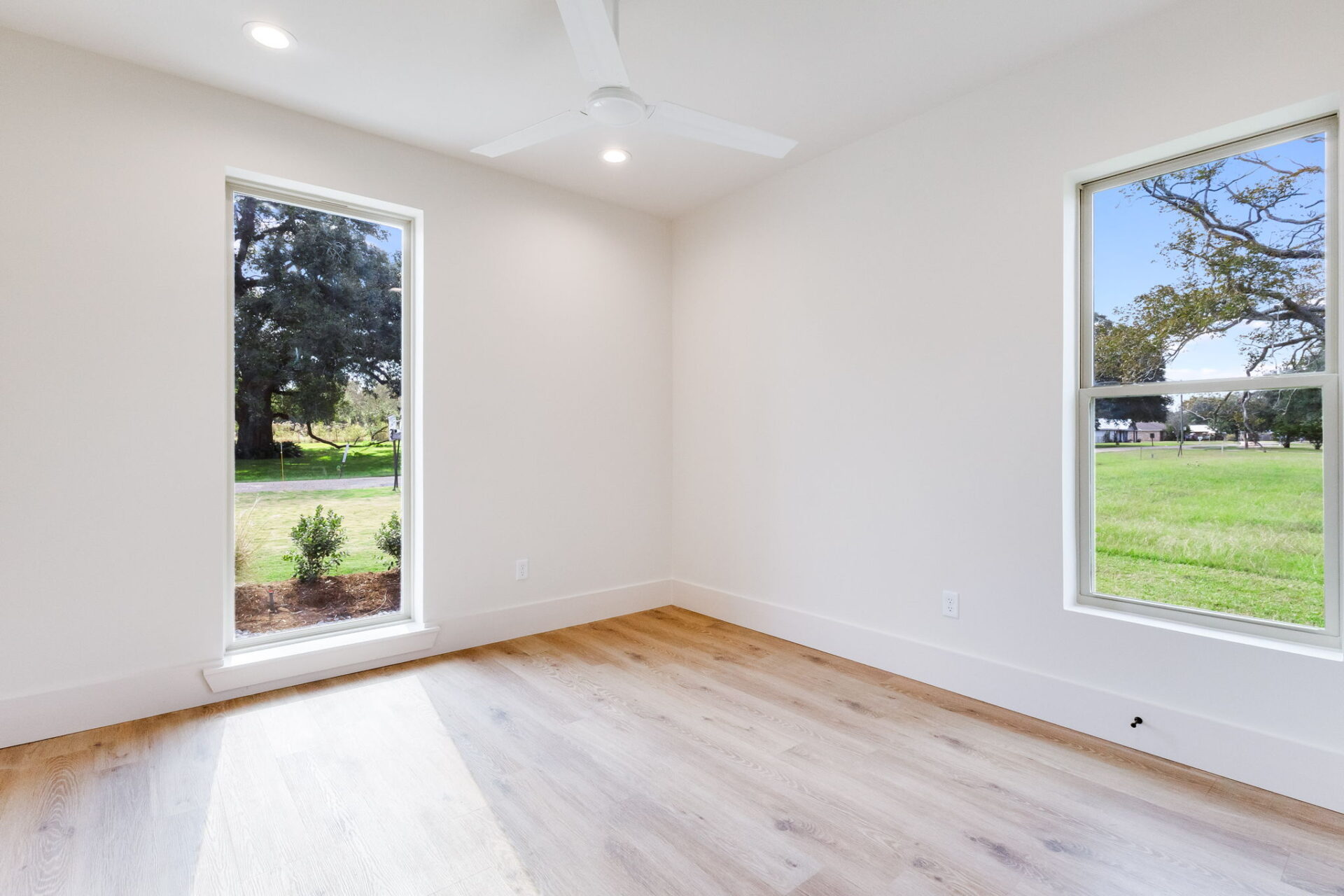 A bright, empty room with light wood flooring, white walls, a ceiling fan, recessed lighting, and two large windows showing a grassy outdoor area with trees.