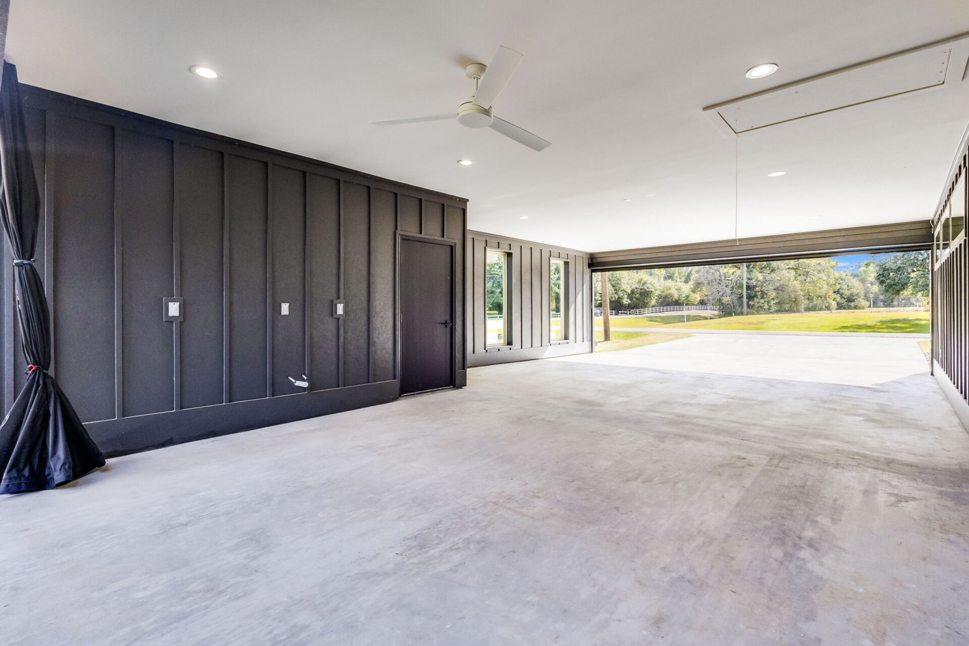 A spacious, empty garage with concrete floors, dark paneled walls, a ceiling fan, ceiling lights, and large open doors leading to a grassy outdoor area with trees in the background.