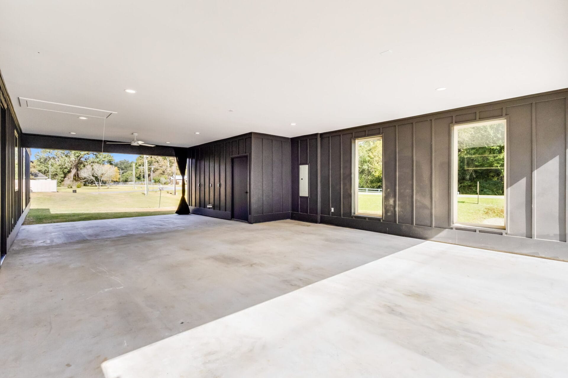 Spacious, empty covered outdoor area with concrete flooring, gray paneled walls, large windows, and an open view to a grassy yard with trees in the background.