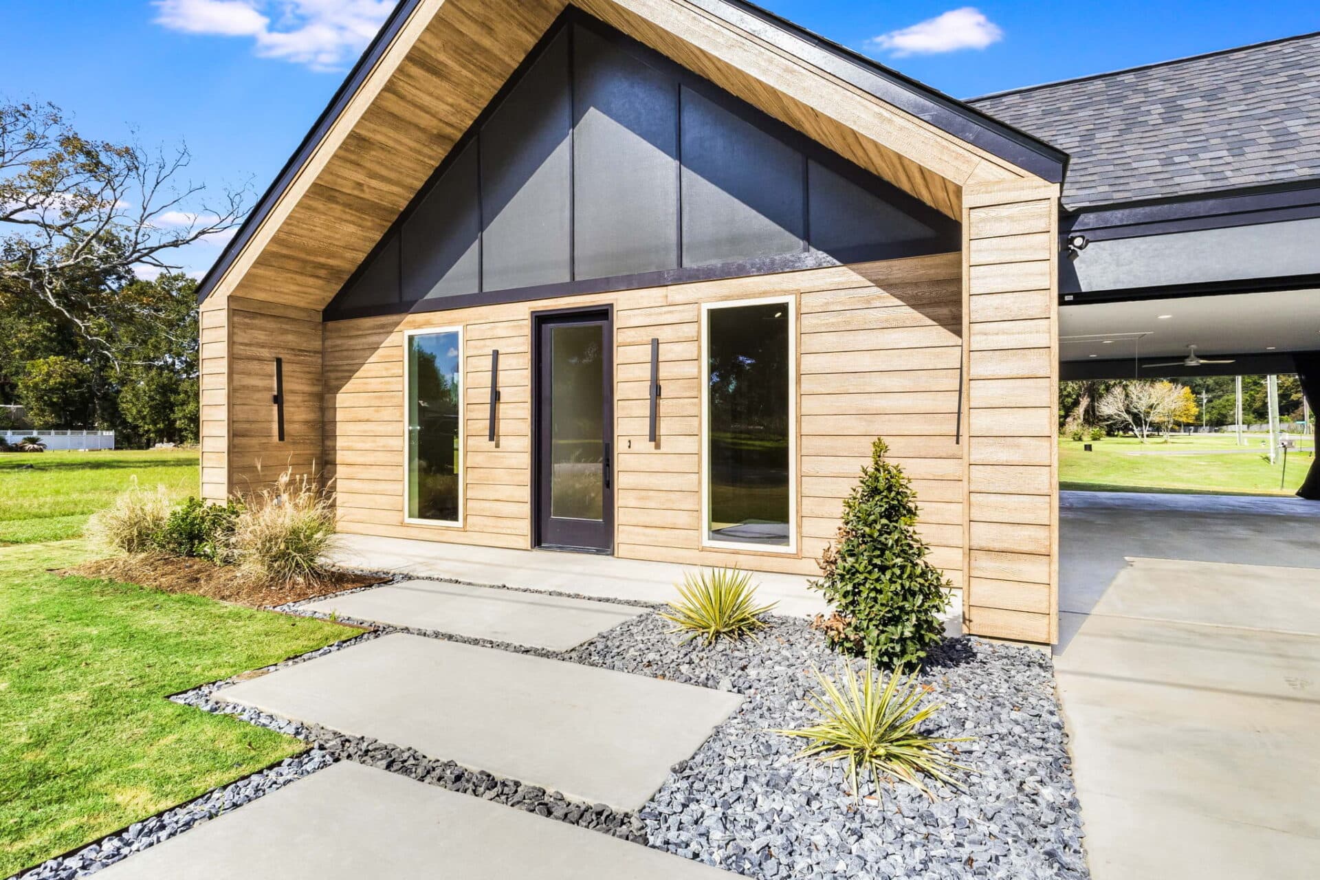 A modern house exterior with light wood siding, large windows, a black front door, and a covered carport. The landscaping includes concrete walkways, small bushes, and decorative rocks.