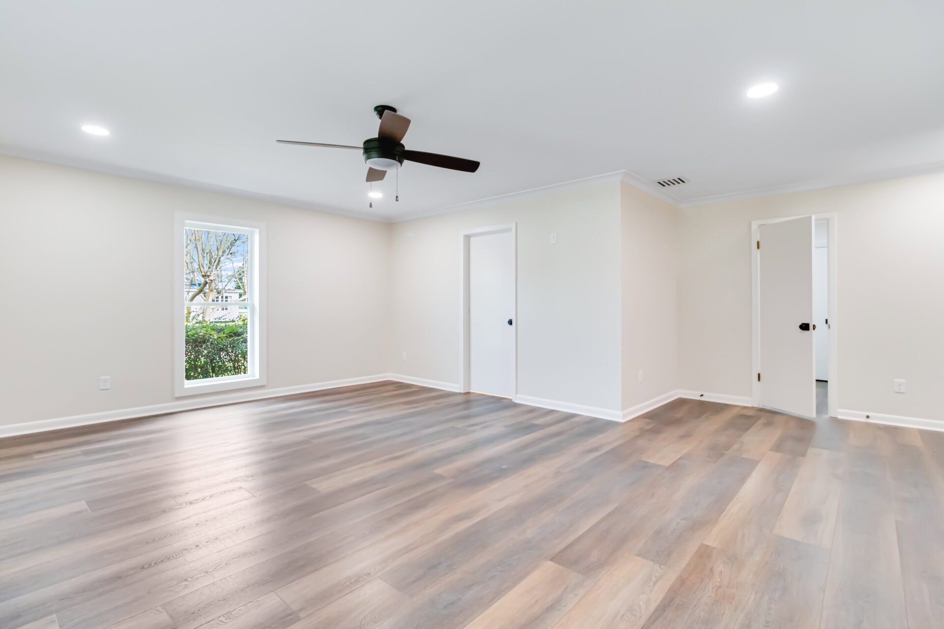 Empty room with light-colored walls, wood-style flooring, a black ceiling fan with light, a window letting in natural light, recessed ceiling lights, and two white doors leading to other spaces.