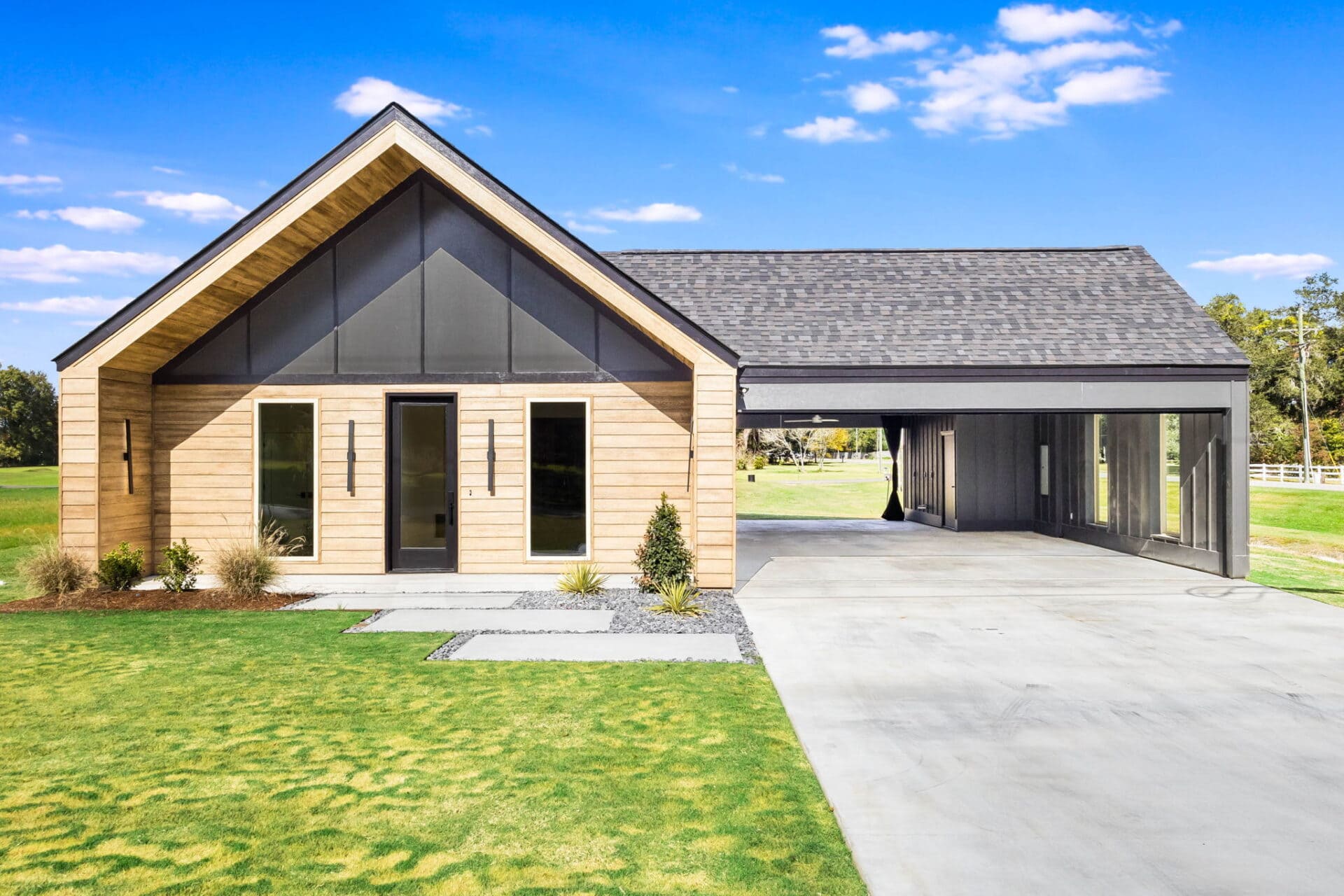 A modern one-story house with light wood siding, large vertical windows, and a black roof, featuring an attached open carport and surrounded by green grass under a blue sky.