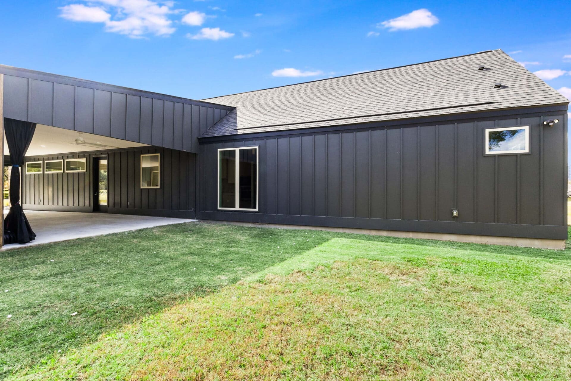 A modern house with dark vertical siding, several windows, and a covered patio area. The yard is mostly green grass, and the sky above is clear with a few scattered clouds.