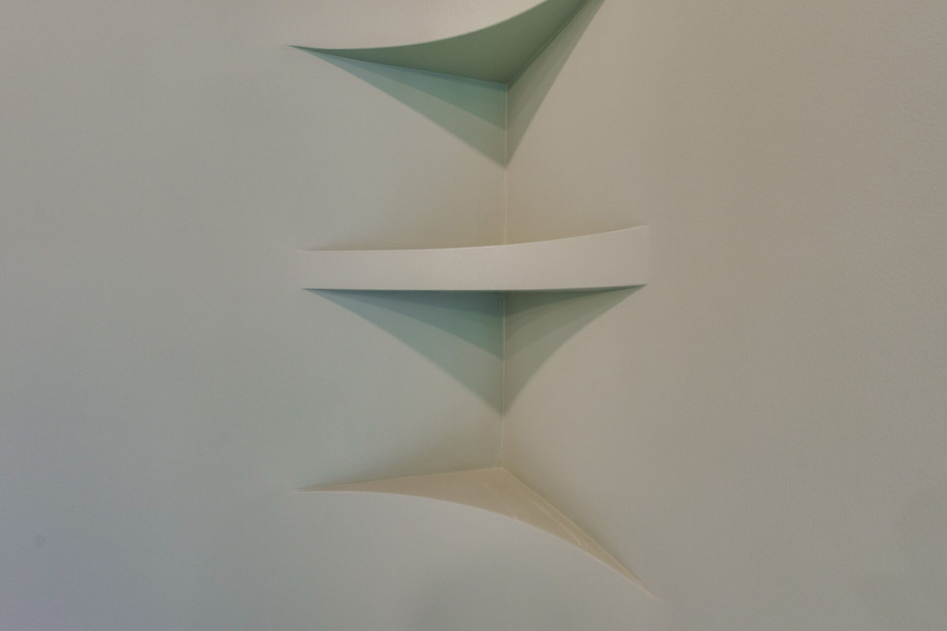 A close-up view of two white, triangular floating shelves mounted in the corner of a light-colored wall, with angled shadows cast on the surfaces below.