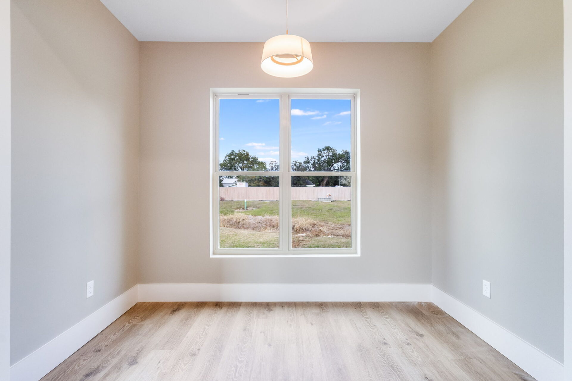 A small, empty room with light-colored walls and wood flooring, featuring a large window in the center that looks out onto a grassy yard with trees and a wooden fence. A ceiling light hangs above.