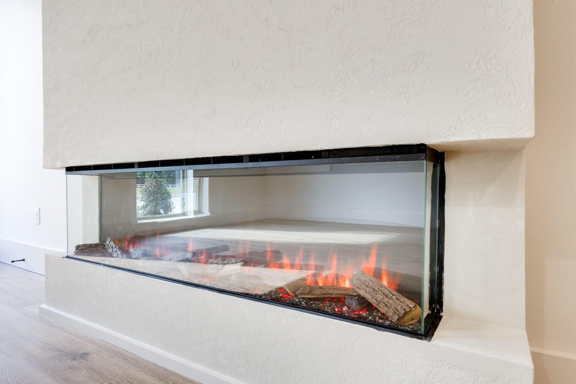 Rectangular built-in electric fireplace with a glass front, displaying artificial logs and orange flames, set into a white textured wall with light wood flooring and a window seen in the background.