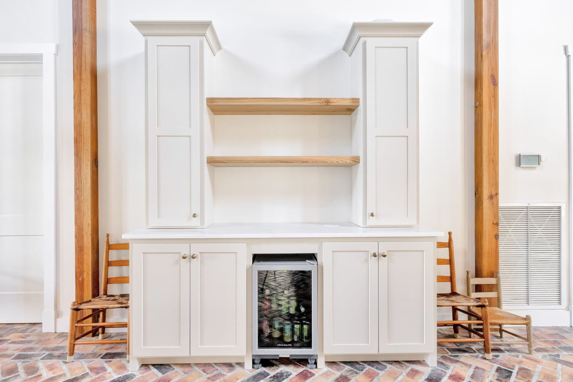 A built-in white cabinet with two open wooden shelves, two tall side cabinets, a countertop, a mini fridge stocked with drinks, and two wooden chairs on either side. The floor is covered in a herringbone brick pattern.