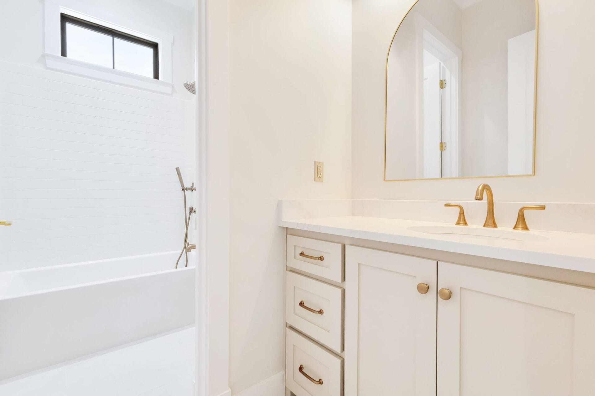 A bathroom with a white vanity featuring gold hardware, a gold faucet, and a large arched mirror. In the background, there is a white bathtub with a showerhead and a window above it.