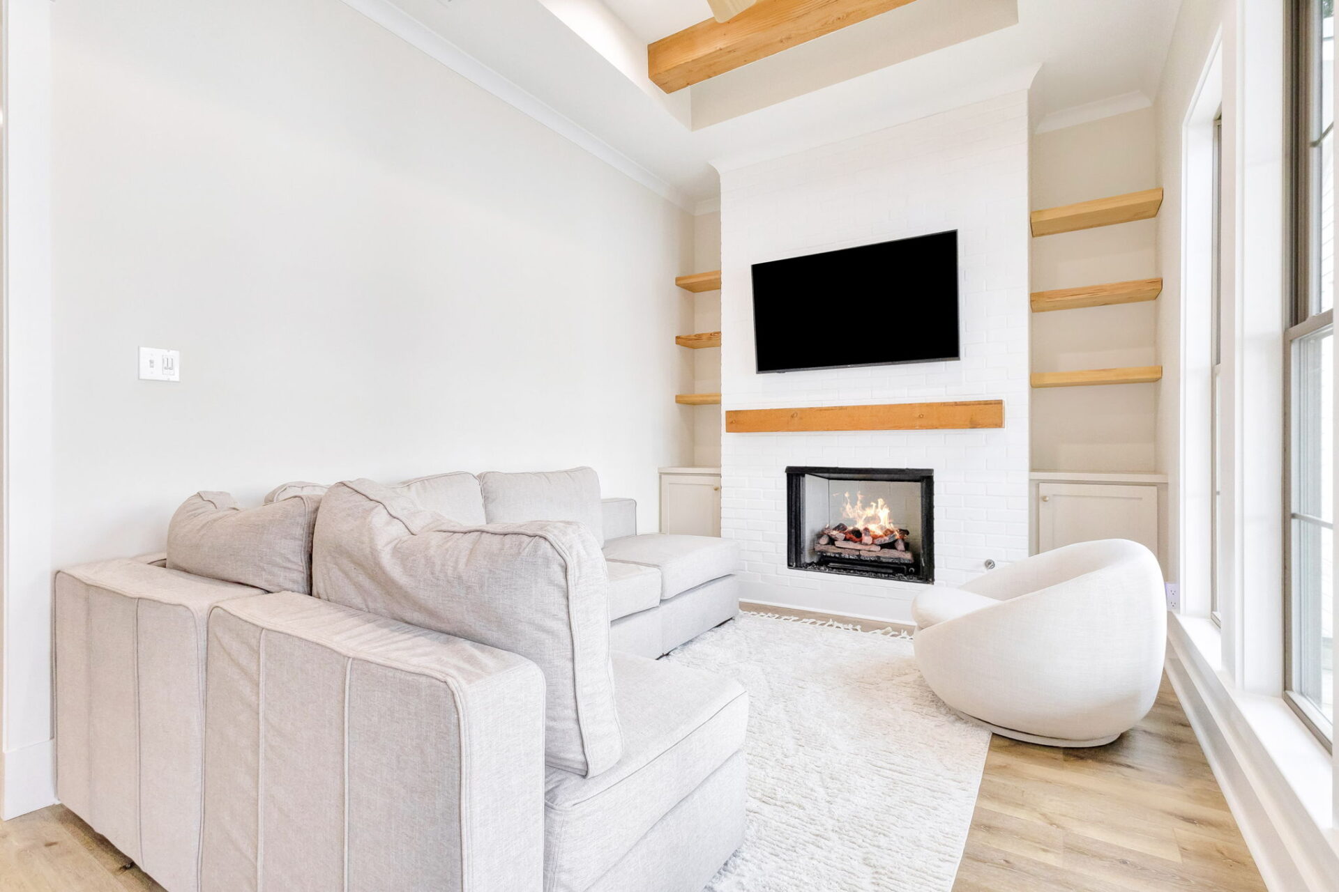 A modern living room with a gray sectional sofa, a round white chair, a fireplace with a wood mantel, a wall-mounted TV, built-in wooden shelves, light wood floors, and large windows letting in natural light.