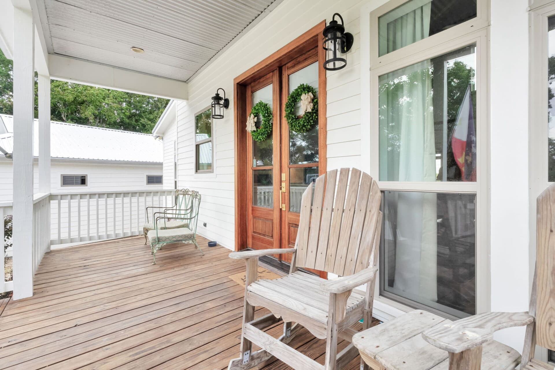 A wooden porch with two Adirondack chairs, a green metal chair, double wooden front doors with wreaths, a lantern-style light fixture, and a large window. Trees and another white building are visible in the background.