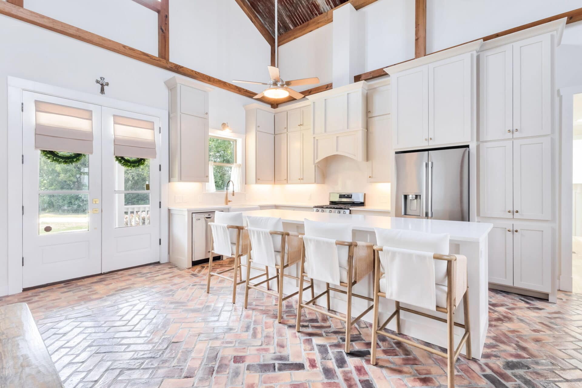 A bright kitchen with white cabinets, stainless steel appliances, and a center island with four wooden chairs. The floor is laid with herringbone-patterned brick, and windows let in natural light.