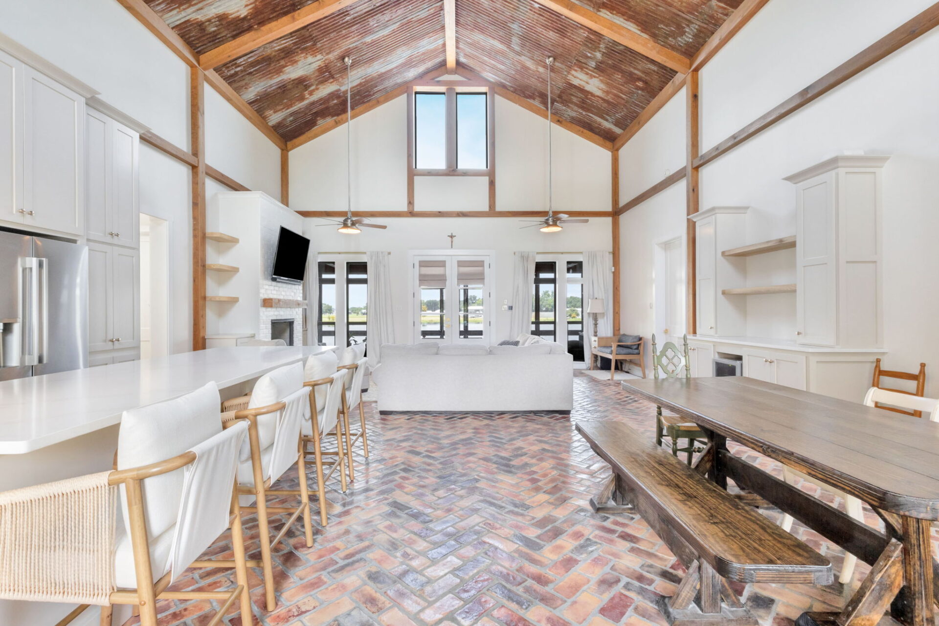 Spacious open-concept kitchen and living area with high vaulted ceilings, exposed wooden beams, brick herringbone floor, white cabinets, a large island with barstools, a rustic dining table, and large windows providing natural light.