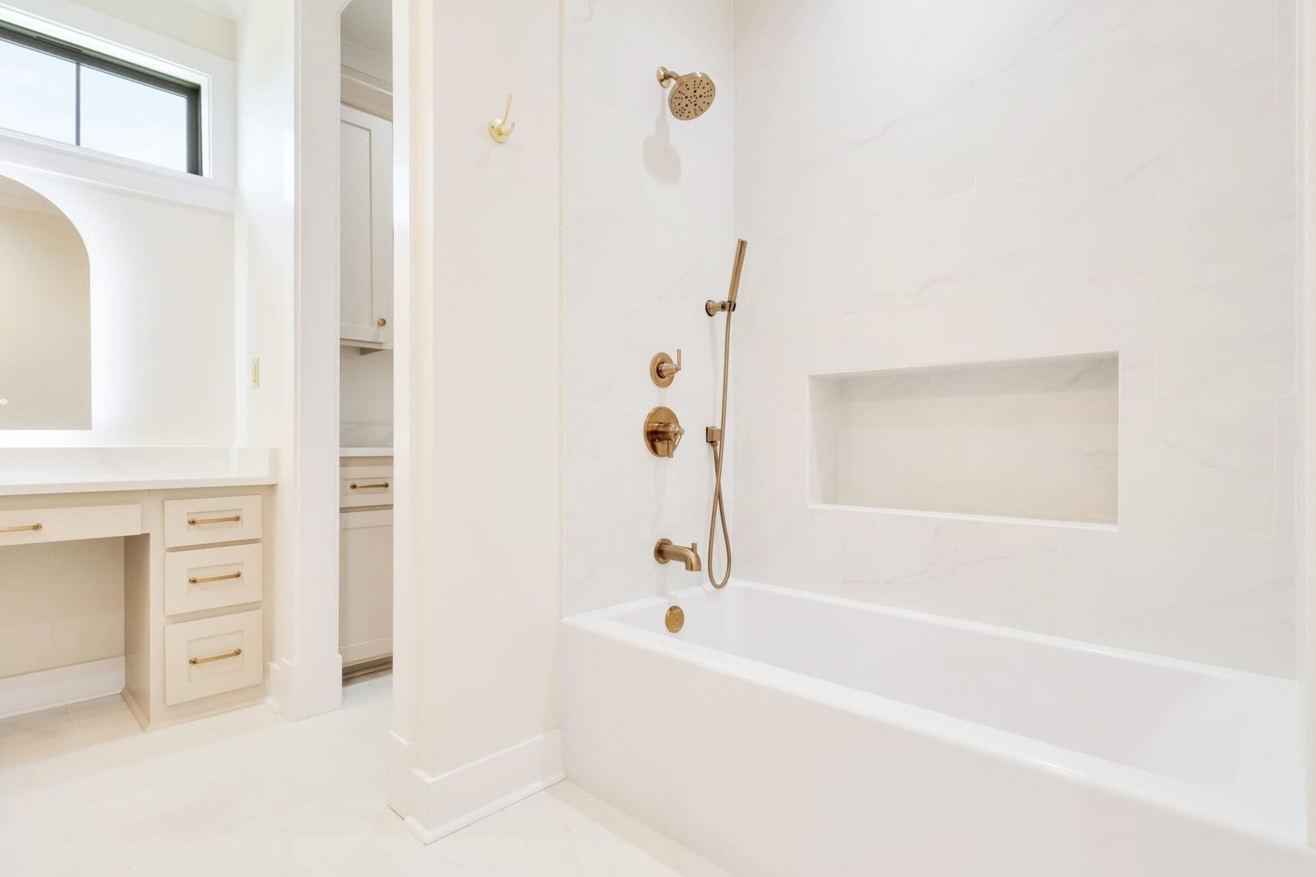 A modern bathroom with a white bathtub, wall niche, and gold shower fixtures. There is a separate vanity area with drawers and a countertop visible on the left side of the image.