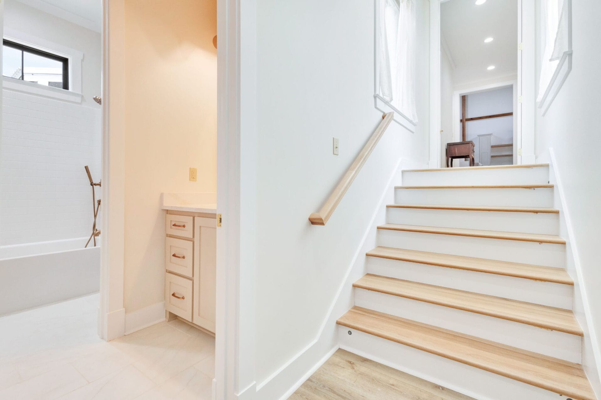 A bright hallway with light wood stairs, a handrail on the right, and a bathroom with a bathtub and white vanity visible to the left. The area is well-lit with natural and ceiling light.