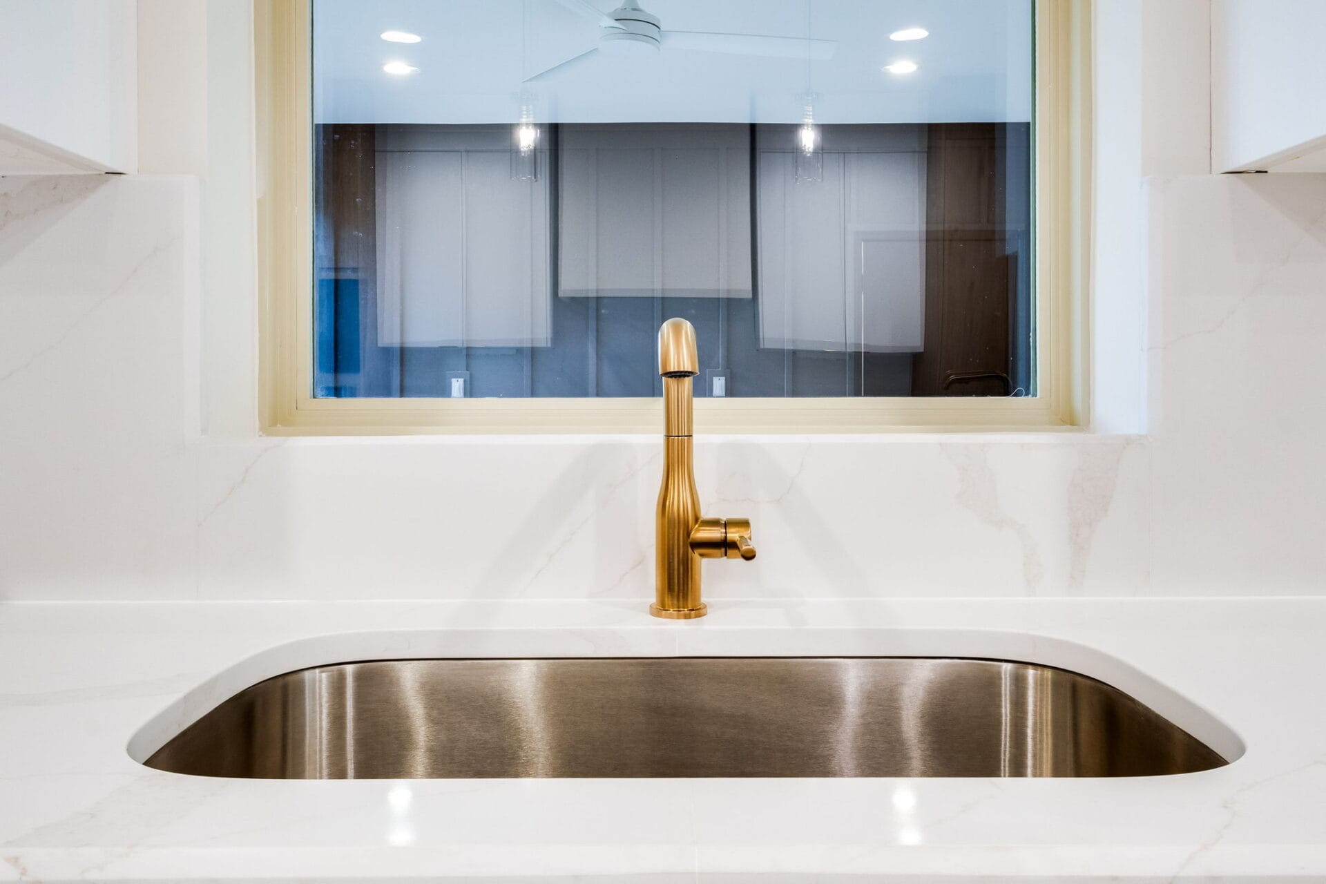 A modern kitchen sink with a gold faucet set in a white countertop, positioned beneath a window. The background shows a reflection of a ceiling fan and cabinets.