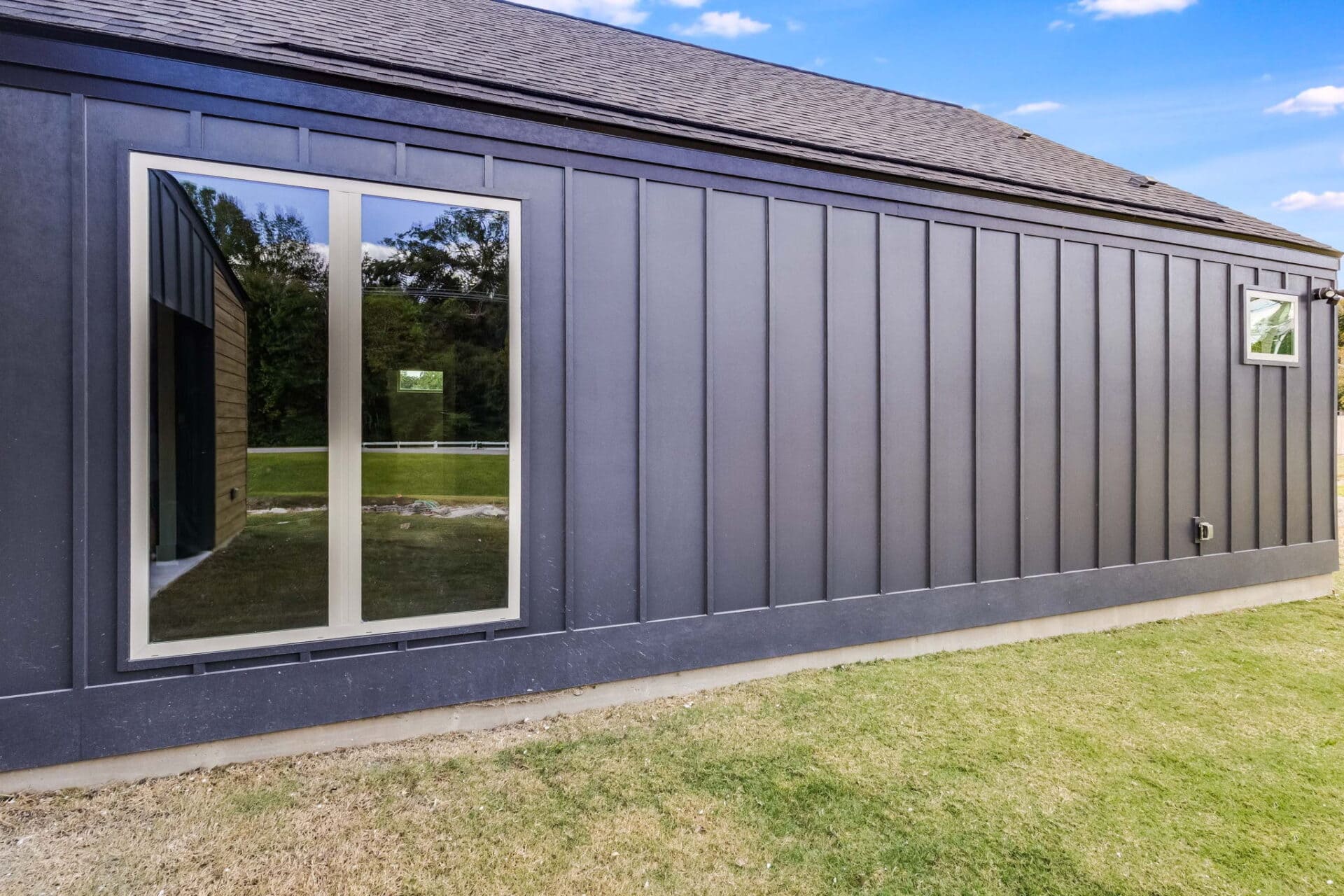 The image shows the exterior wall of a modern house with dark vertical siding, two windows, and a sloped roof, next to a lawn with green grass and trees reflected in the large window.