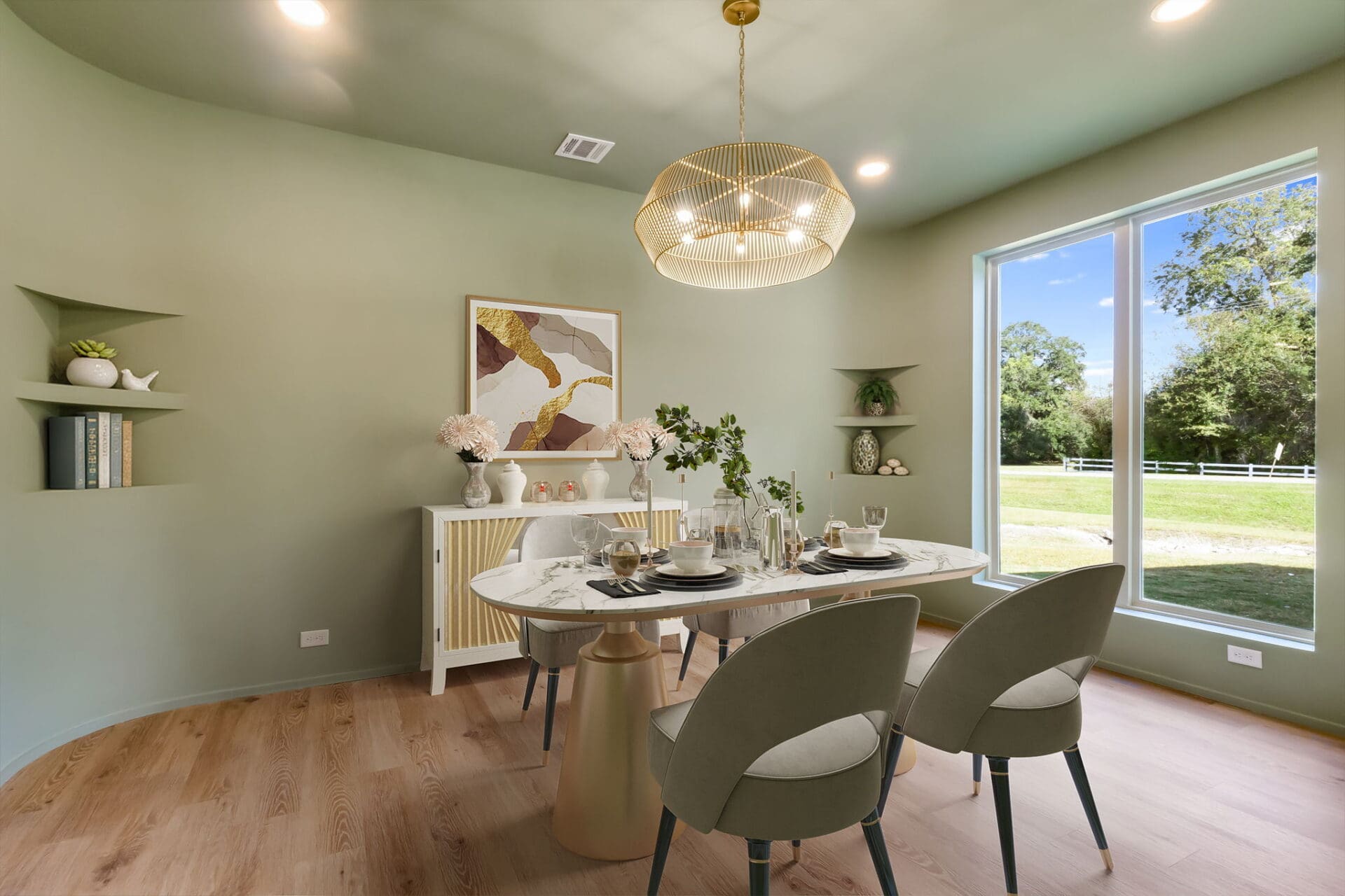 A modern dining room with sage green walls, wood flooring, a round marble table set for four, green chairs, a gold light fixture, large window, wall shelves, and framed abstract art above a buffet.