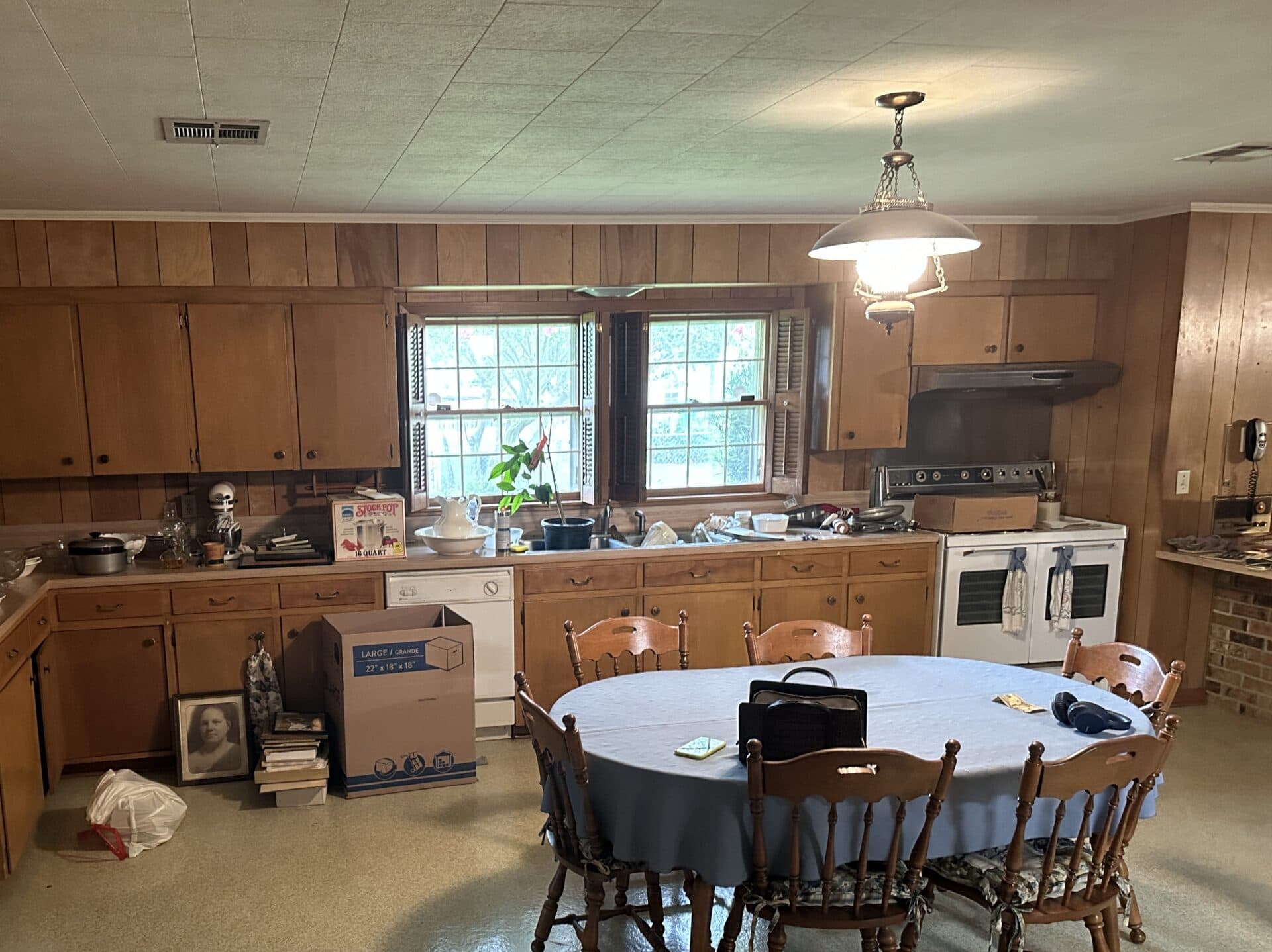 A kitchen with wooden cabinets, a table with chairs and a blue tablecloth, moving boxes, assorted kitchen items on the counters, a stove, and sunlight coming in through two large windows.