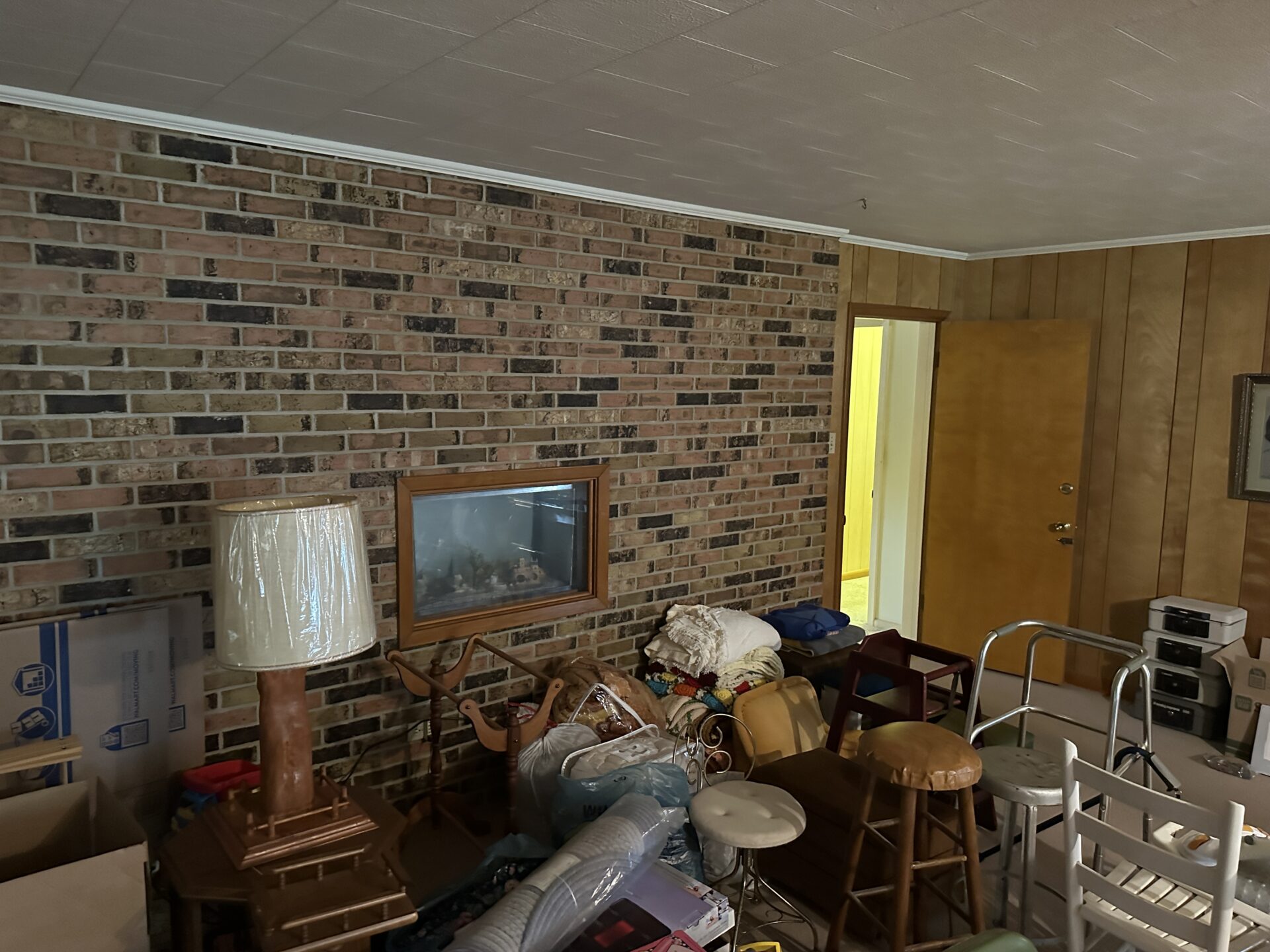 A cluttered room with a brick accent wall, wood paneling, and a closed wooden door. Various furniture, lamps, boxes, and miscellaneous items are scattered around, with a lit hallway visible through an open doorway.