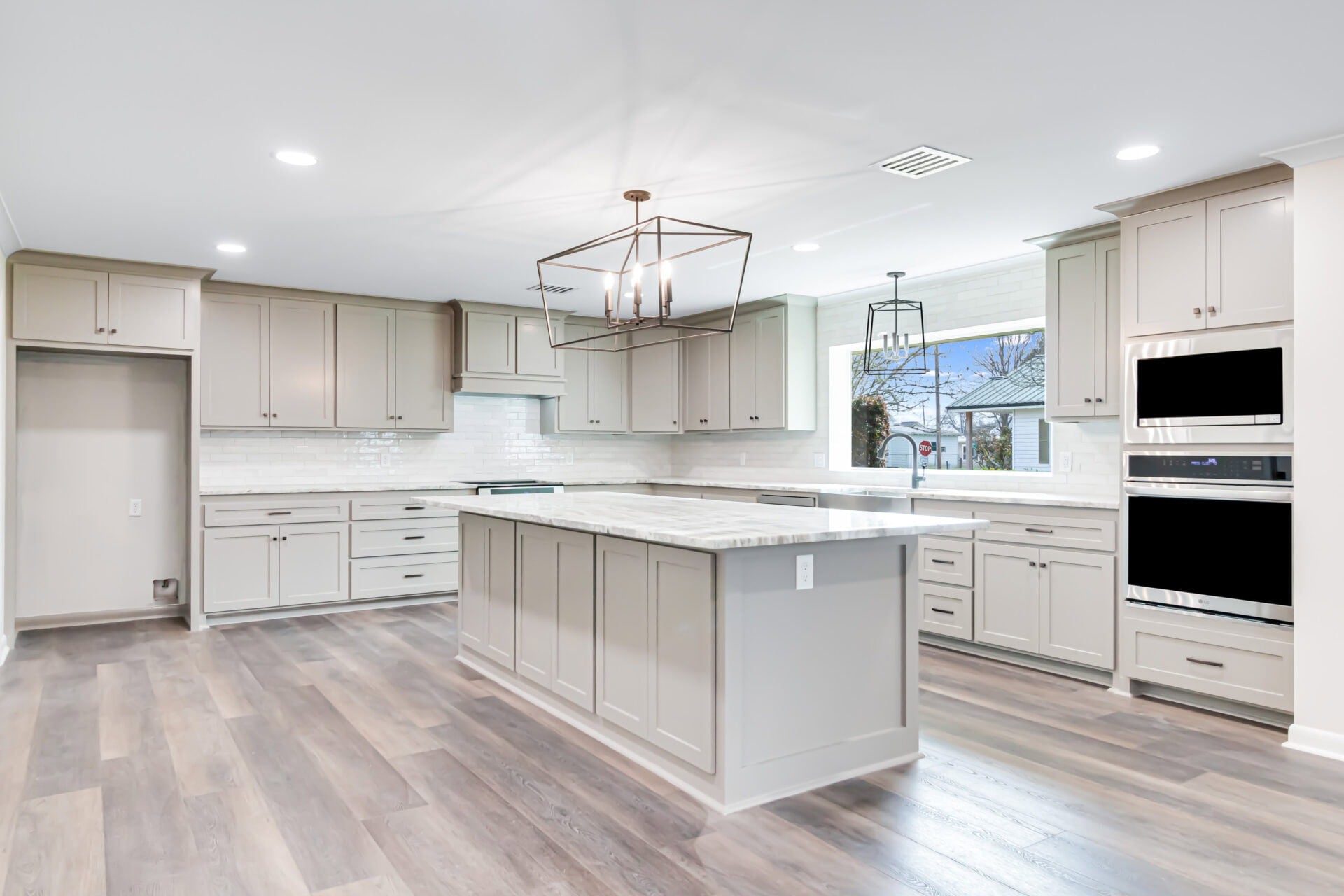 A modern kitchen with light gray cabinets, a large central island, stainless steel appliances, two built-in ovens, white subway tile backsplash, and wide plank wood flooring under bright ceiling lights.