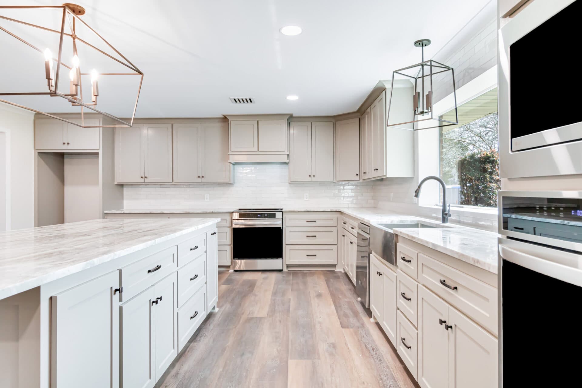 Modern kitchen with white cabinets, stainless steel appliances, marble countertops, a large island, pendant lighting, and wood flooring. White subway tile backsplash and large window over the sink provide a bright, open atmosphere.