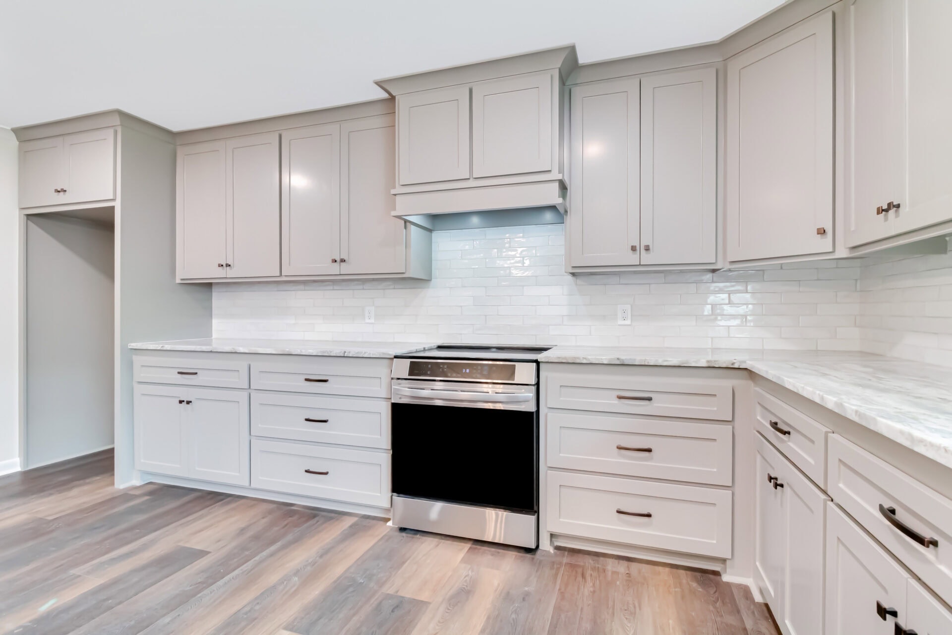 Modern kitchen with light gray cabinets, white subway tile backsplash, stainless steel oven, and marble countertops. The floor is wood with a medium to dark finish. There are no visible appliances or decorations.