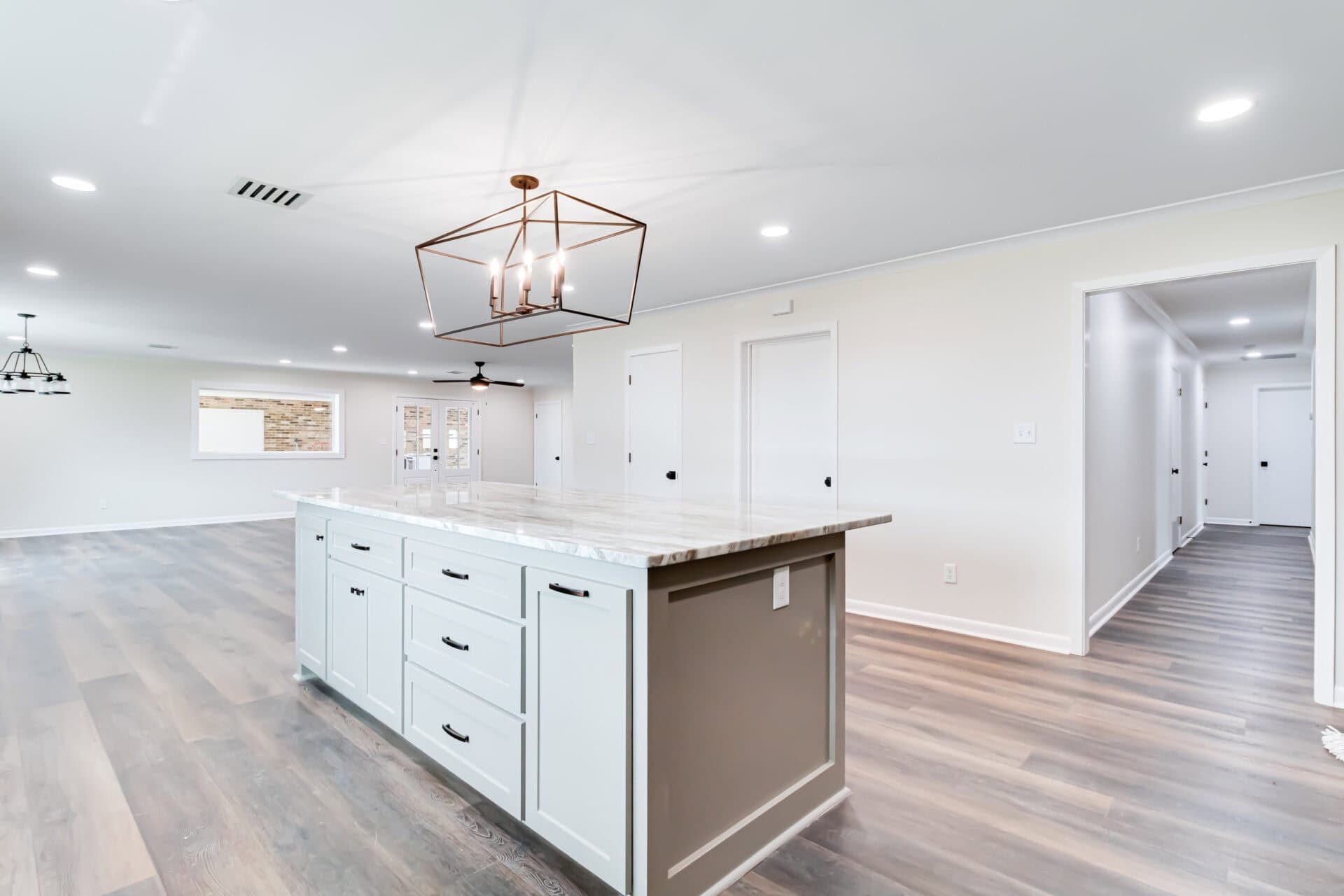 Modern kitchen with light gray walls, wood flooring, and a large marble-topped island with drawers and cabinets. A geometric light fixture hangs above the island, and hallways are visible in the background.