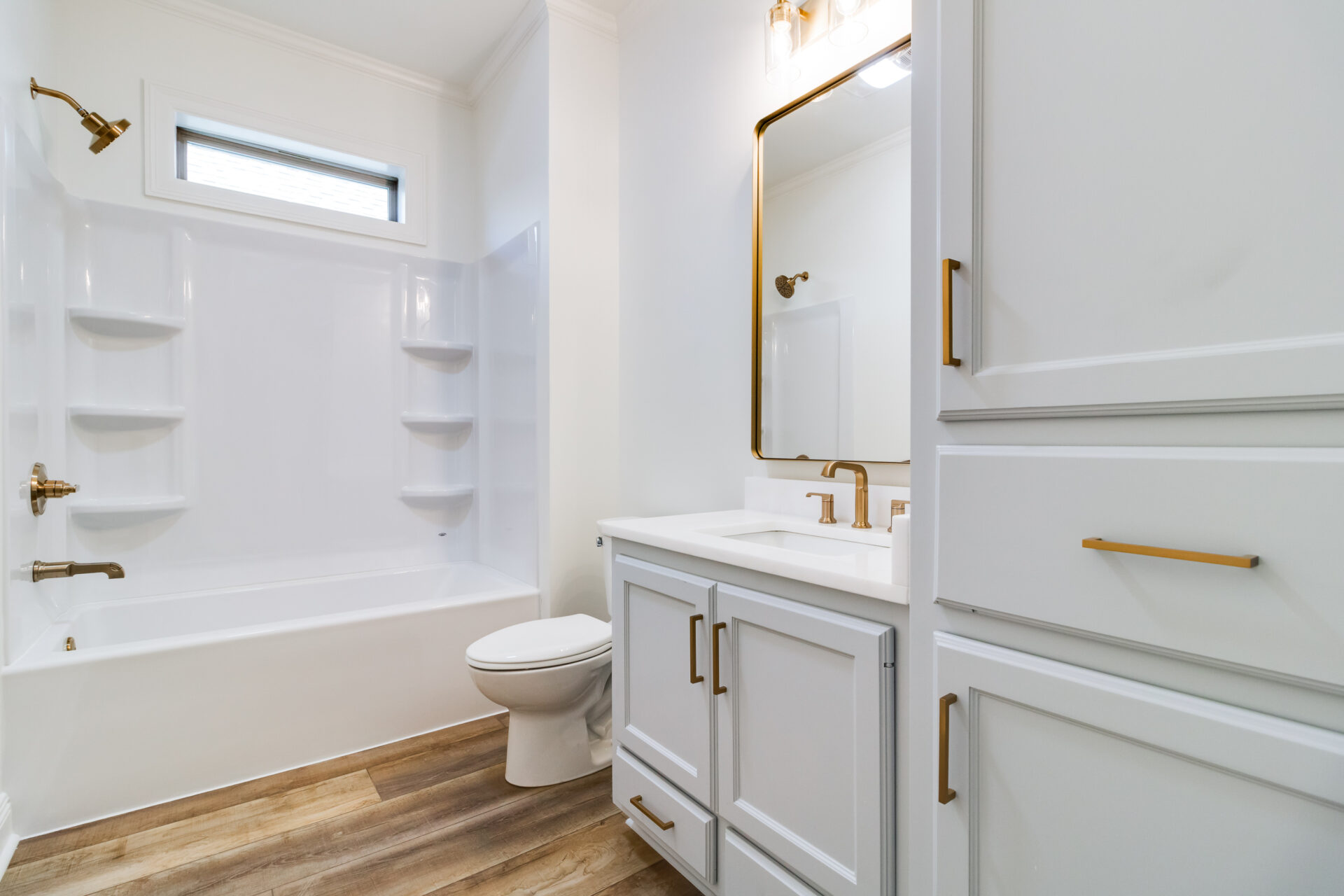 A modern bathroom by Hebert Reno with white walls and fixtures. It features a bathtub with built-in shelves, a showerhead, a toilet, a vanity with a rectangular sink, a large mirror, and a cabinet. The floor has wood-style tiles, and gold accents are present on faucets and handles.