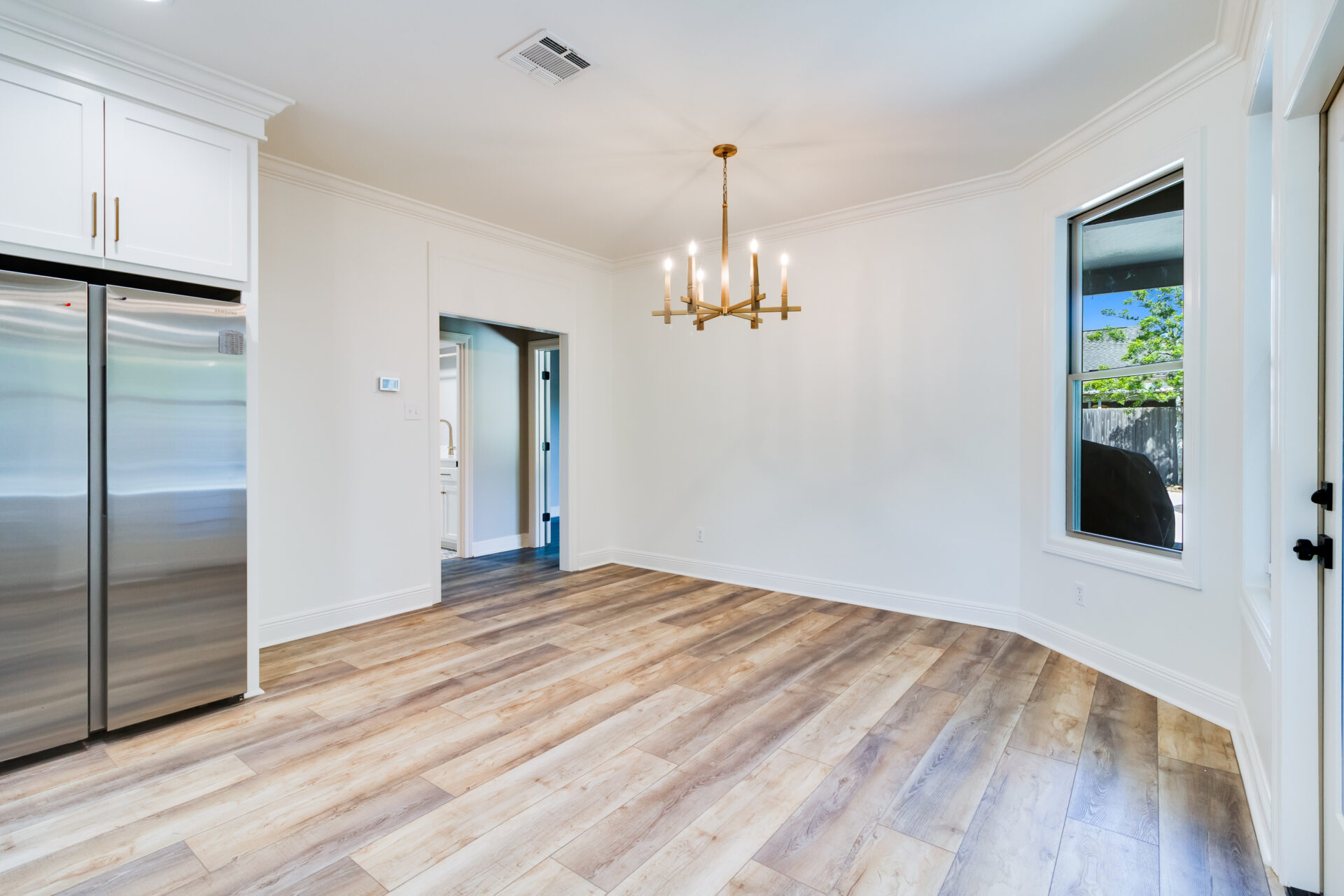 A modern, unfurnished dining area with light wood flooring and white walls by Hebert Reno. The room features a large window, a gold-colored chandelier, and an adjacent kitchen area visible through a doorway. Stainless steel appliances are also seen in the kitchen.