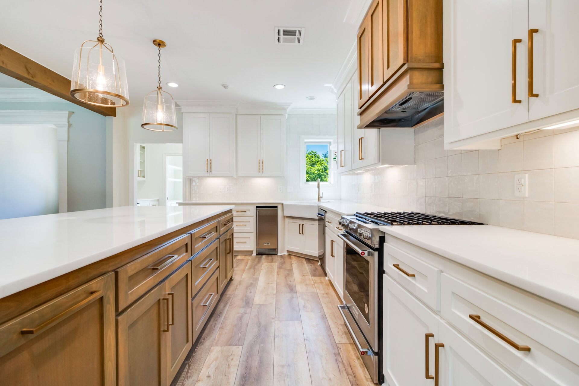 A modern kitchen by Hebert featuring wooden and white cabinetry, a large island with a white countertop, stainless steel appliances, and pendant lights. The light wood floor complements the design, while a window allows natural light to enter, making this Reno space both functional and beautiful.
