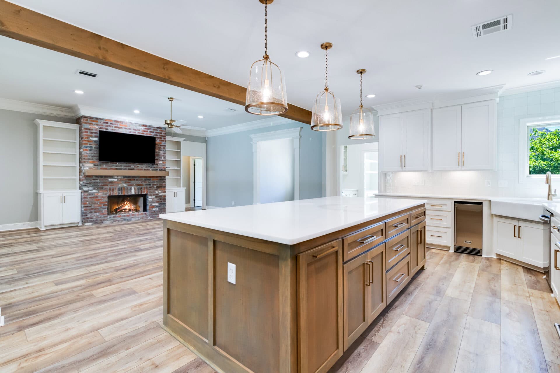 A spacious modern kitchen with white cabinets and an island featuring a wooden base and white countertops, crafted by Hebert Reno. Pendant lights hang above the island, illuminating the space. The open layout extends to a living area with a brick fireplace and built-in shelves.