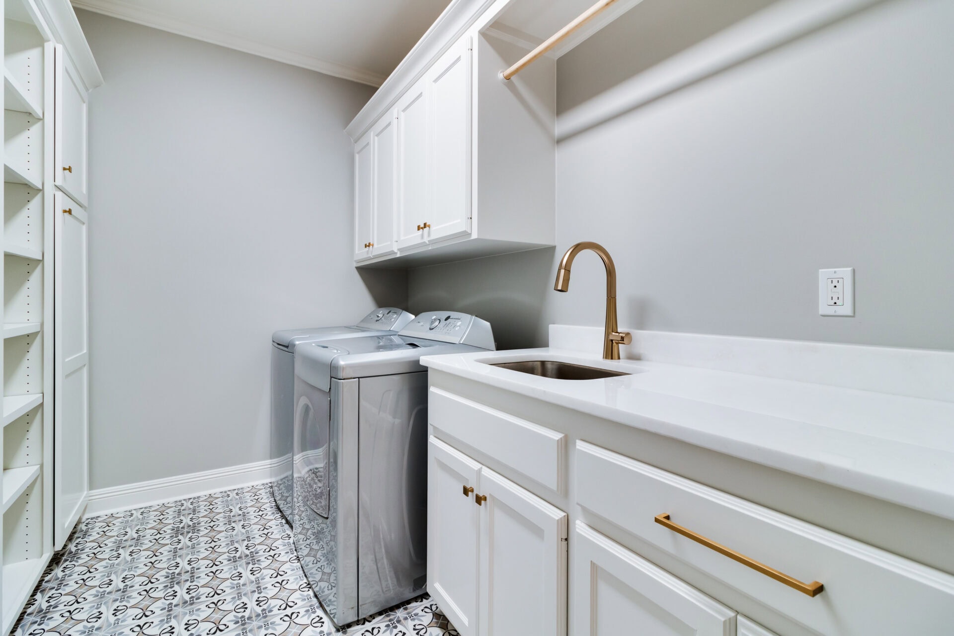 A modern laundry room by Hebert Reno features white cabinetry, a stainless steel washer and dryer, a sink with a brass faucet, and a decorative tile floor. Ample cabinets and shelves provide storage, while a hanging rod facilitates drying clothes. The walls are painted light gray for a clean finish.