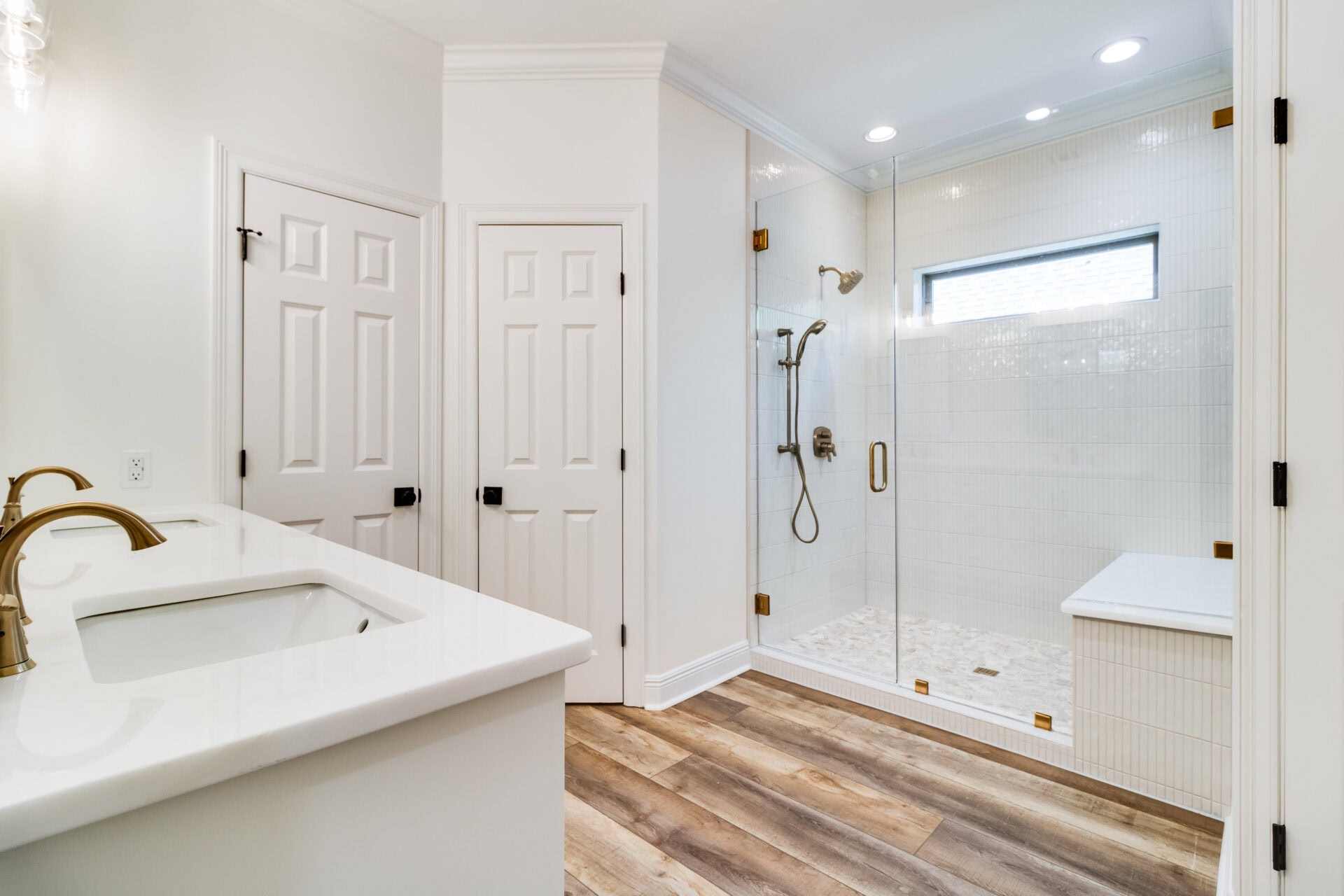 A modern bathroom by Hebert Reno features white walls and fixtures. The space includes a double sink vanity with brass faucets on the left, two doors in the center, and a large glass-enclosed shower with white tiles and multiple showerheads on the right. The floor is finished in elegant wood.