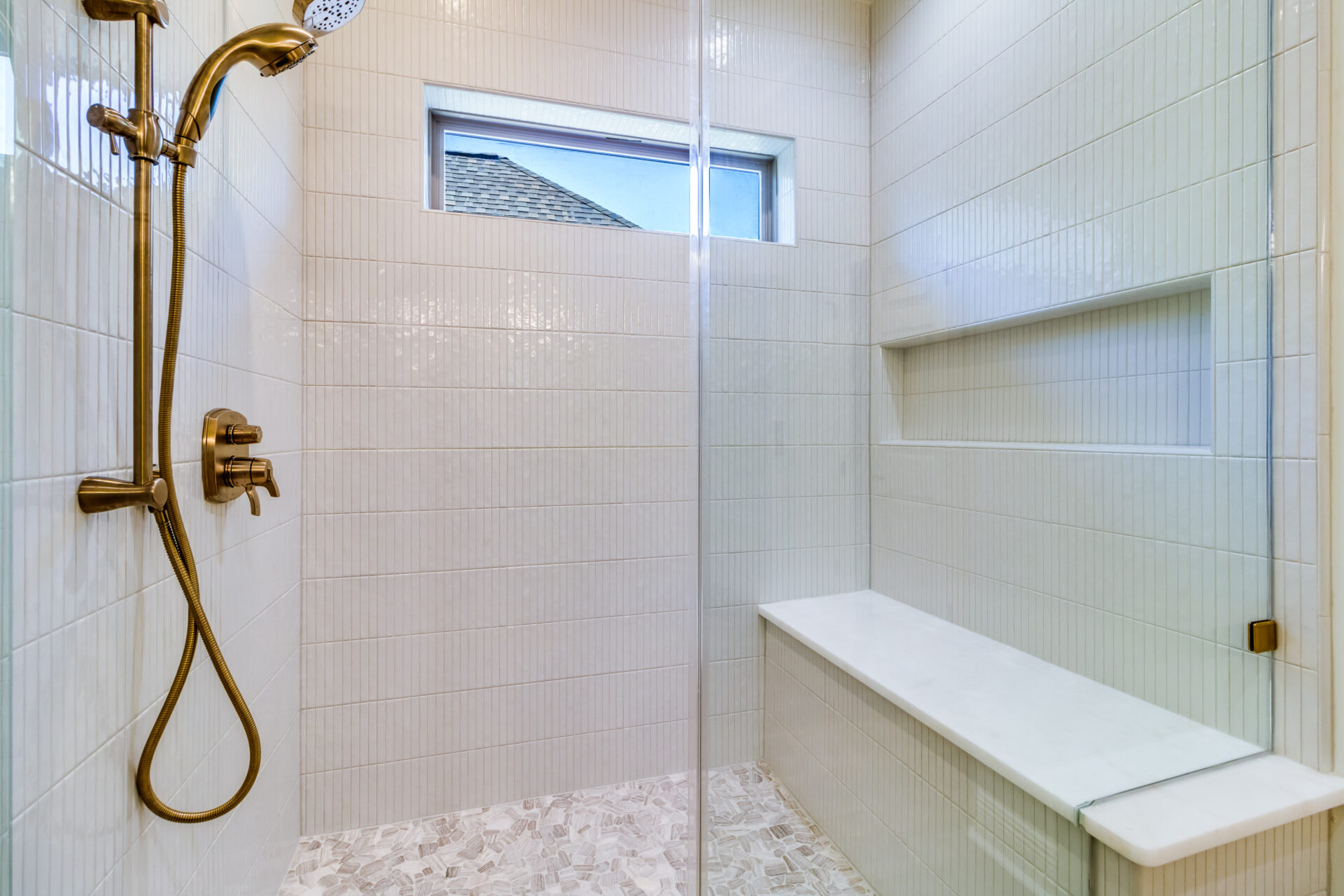 A modern, minimalist shower in the Hebert Reno project features light-colored tiles, a glass door, and brushed gold fixtures including a rainfall showerhead and hand-held shower. The shower has a built-in bench and a high window letting in natural light.