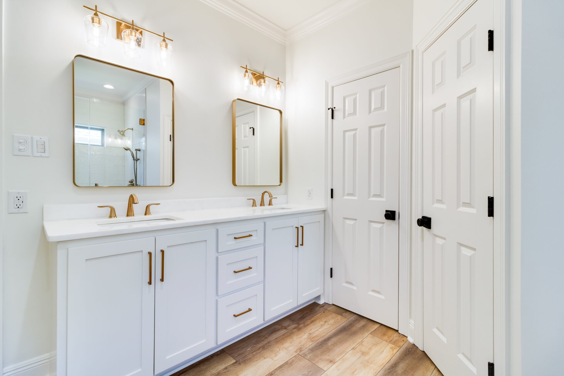 A Hebert Reno bathroom showcases a double vanity with two square mirrors and brass fixtures. The white cabinets, adorned with gold handles, are complemented by a sleek white countertop. With two closed doors and wooden flooring, the light-colored walls complete this elegant space.